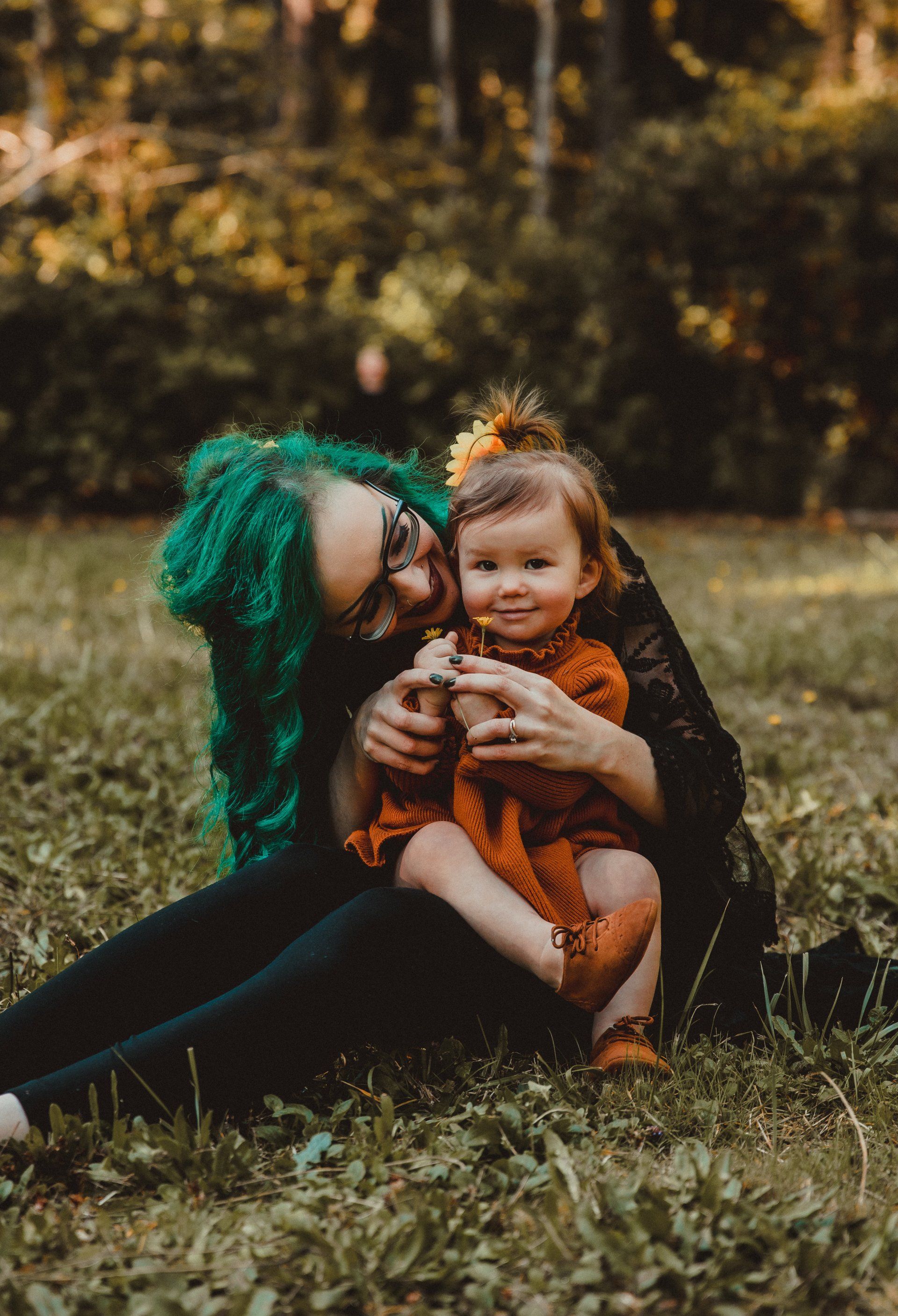 Woman with green hair and a child on a grassy lawn. The woman is smiling, holding the child. The child holds a flower.