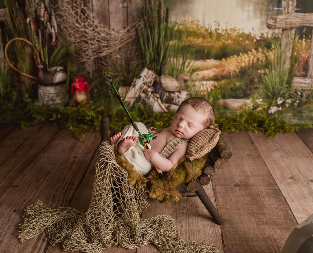 Newborn baby asleep in tiny fishing chair with fishing rod. Rustic setting with netting and greenery.