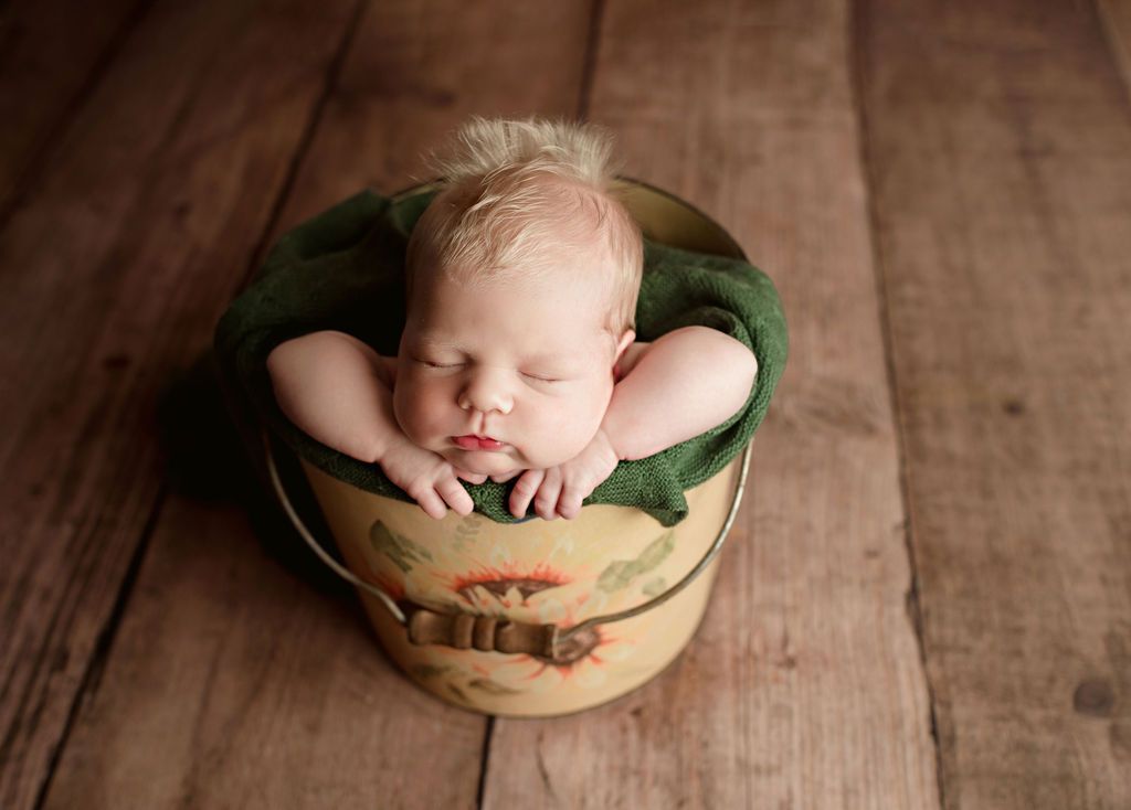 Sleeping baby in a green blanket inside a vintage painted bucket on a wooden floor.
