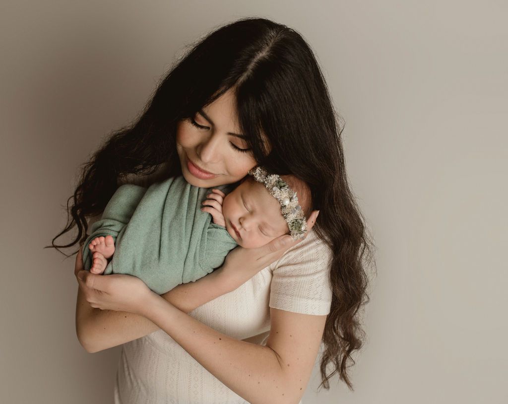 Woman holding a newborn baby. The baby sleeps, wearing a floral headpiece, and is wrapped in a green cloth.