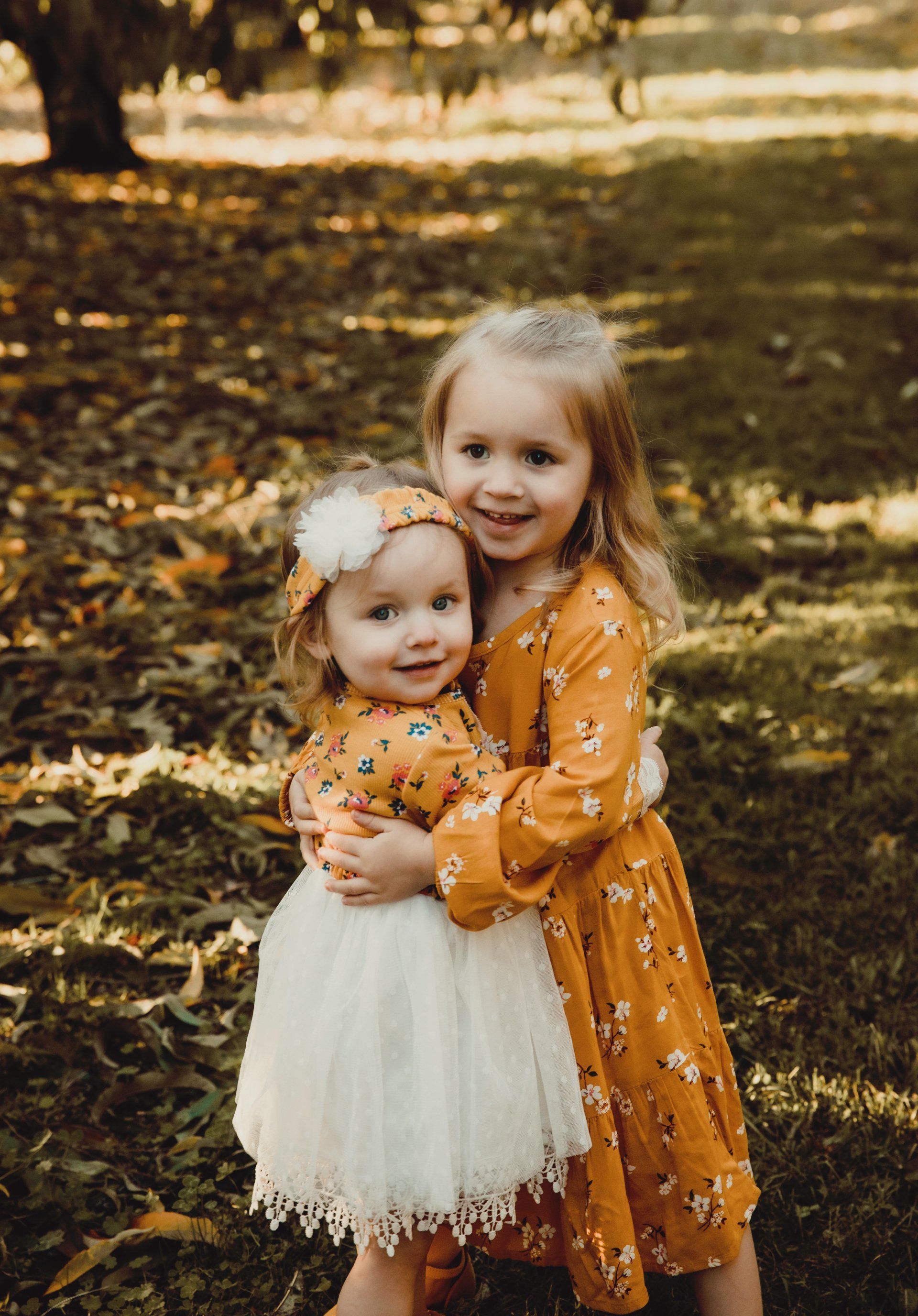 Two young girls embrace outdoors; fall foliage in background. One wears floral dress, the other a white tutu.