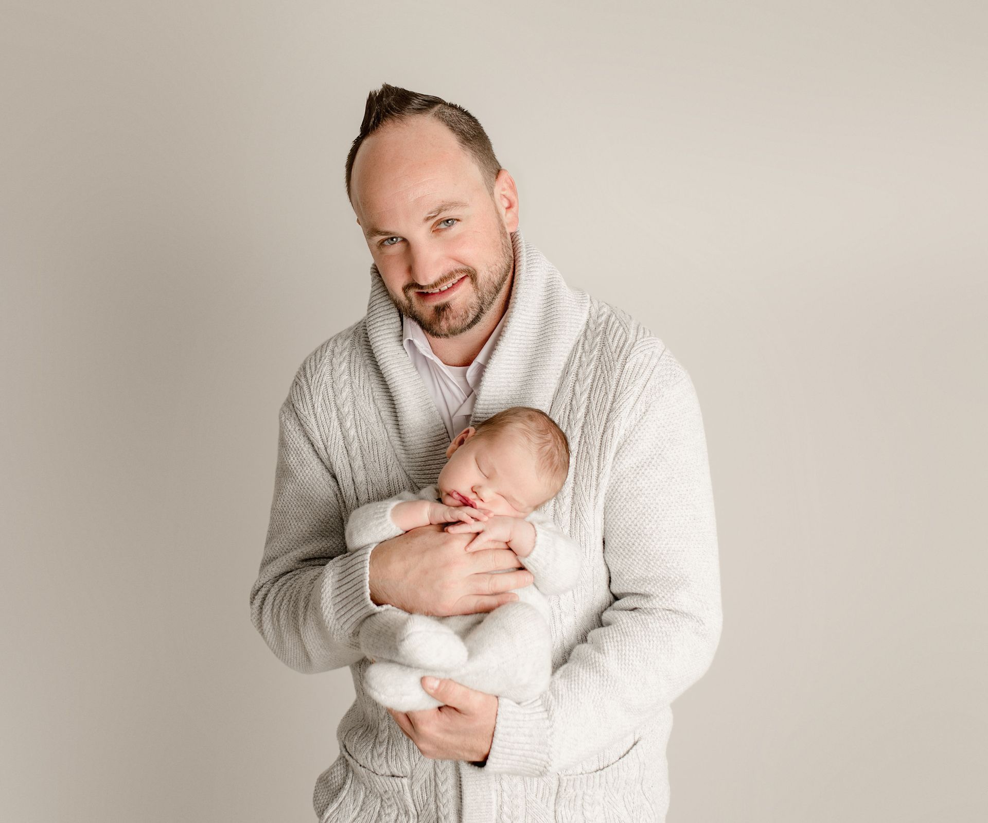 Man in a cable-knit sweater holding a sleeping newborn baby against a neutral background.