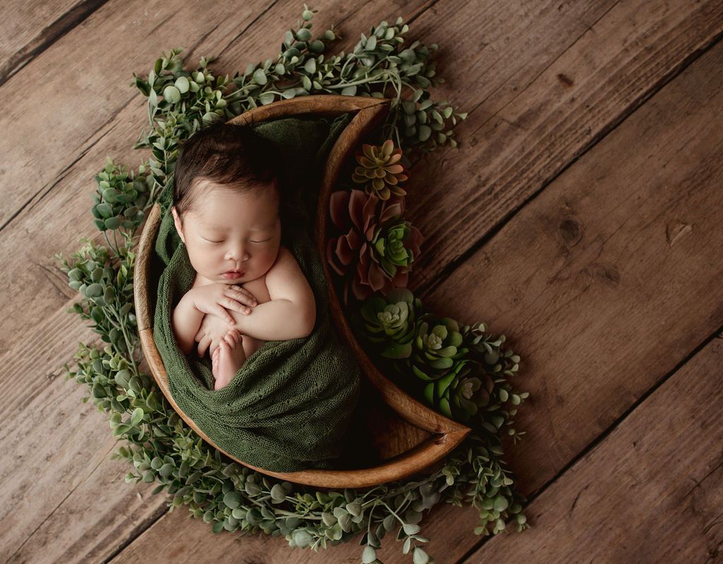 Newborn swaddled in green fabric, sleeping in a crescent moon-shaped prop, surrounded by greenery on a wood background.