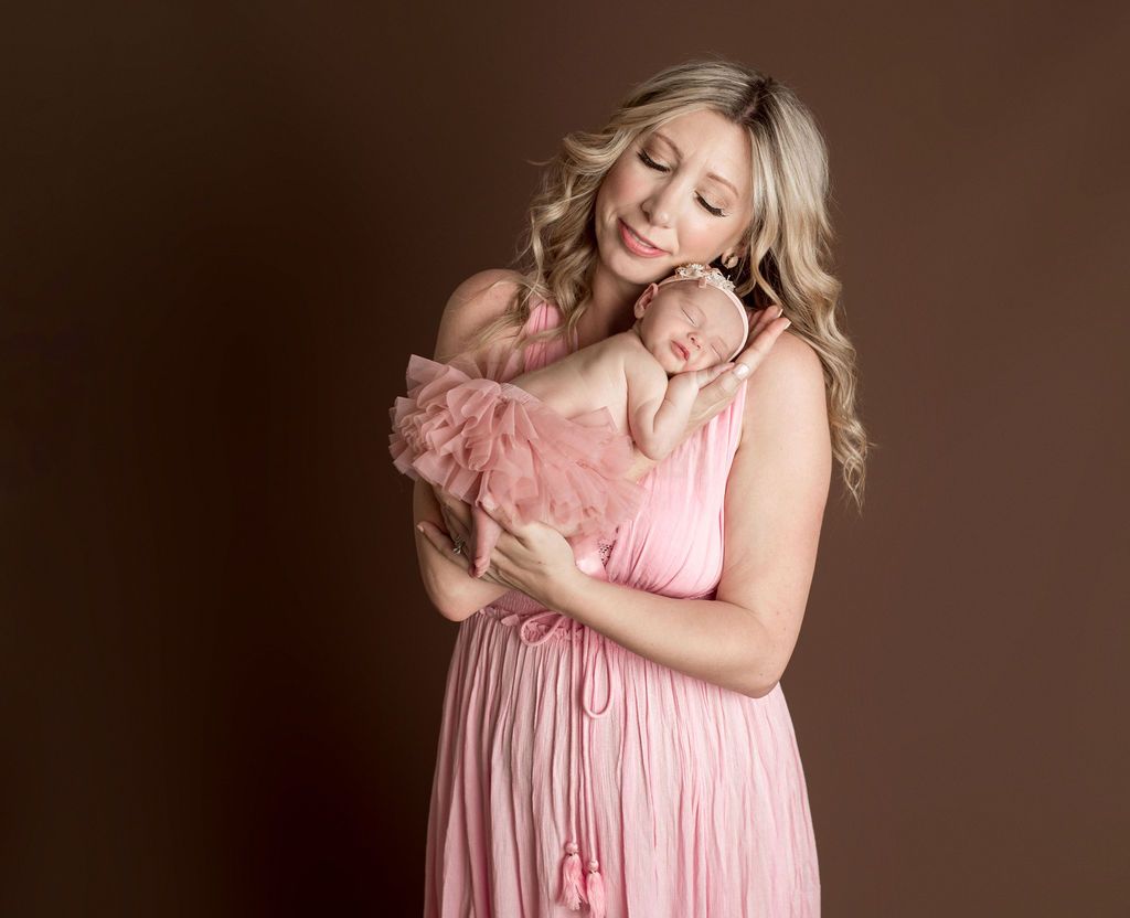 Woman in pink dress holds sleeping baby in pink tutu. Brown backdrop.