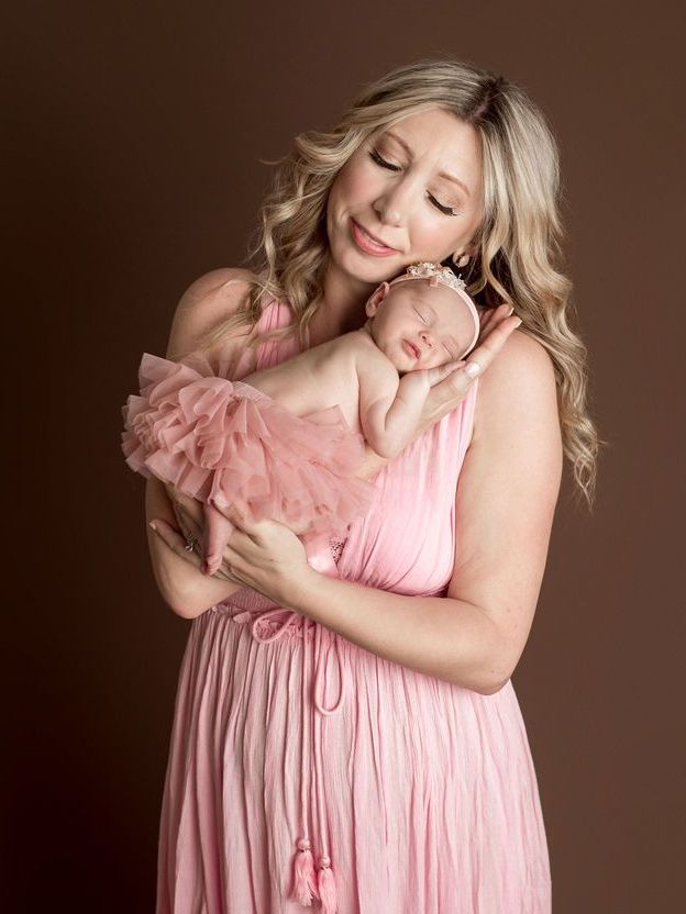 Woman in pink dress holds sleeping baby in pink tutu, brown background.