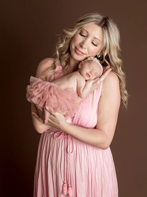 Woman in pink dress holds sleeping baby in pink tutu, brown background.