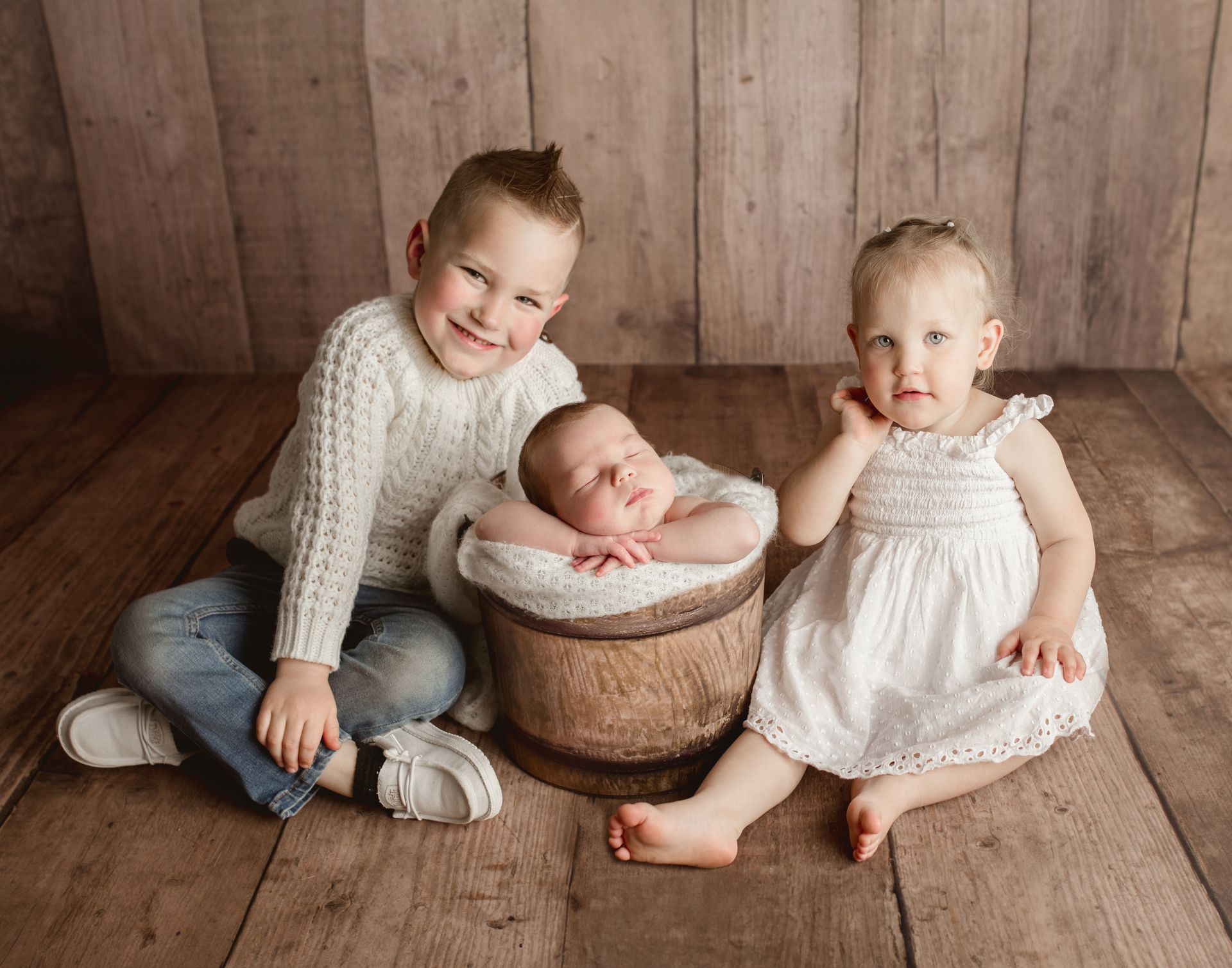 Three children, a baby in a basket, pose on a wood floor. Older child smiles, girl looks curious.