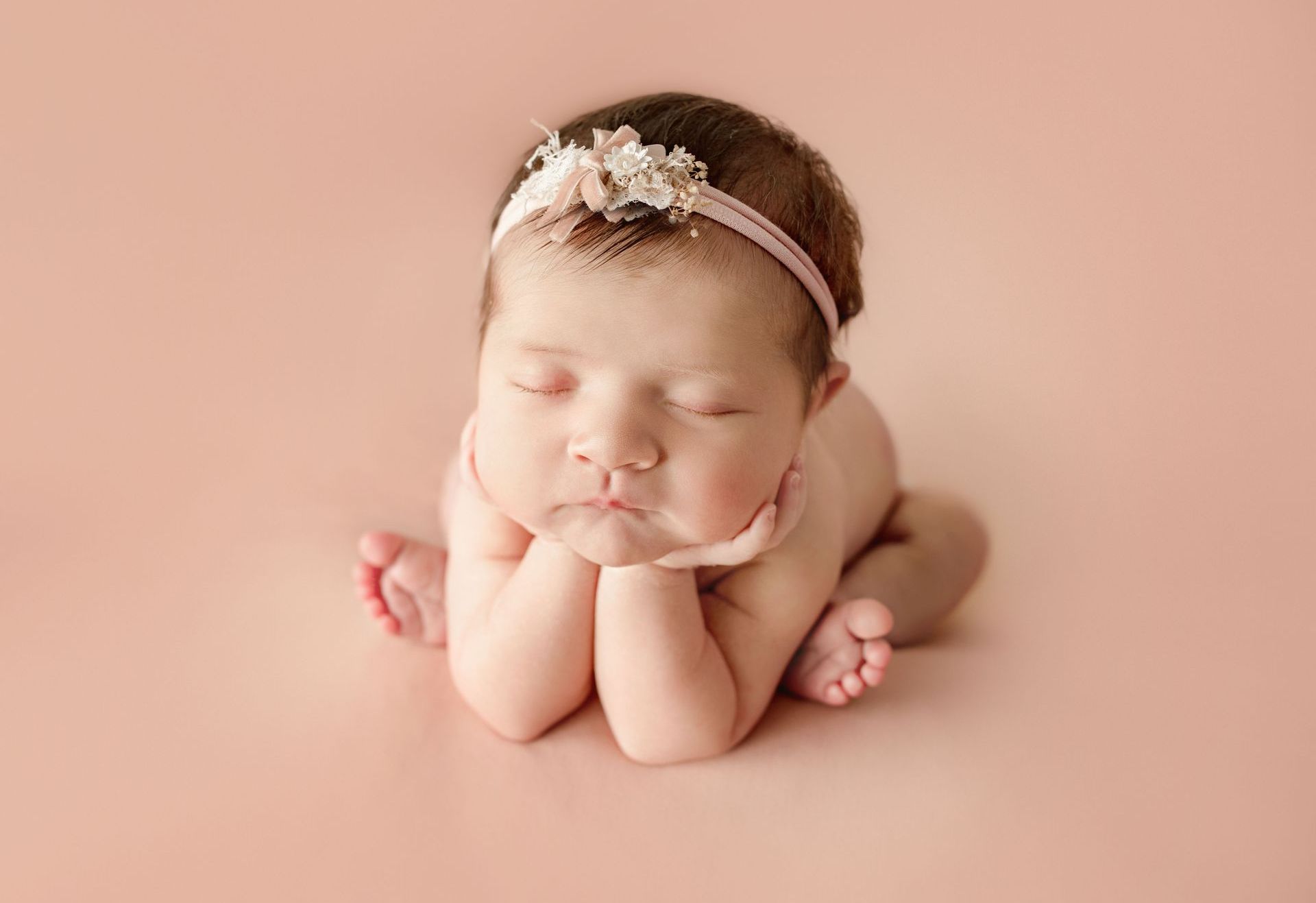 Newborn baby with eyes closed, resting face on hands, wearing a floral headband on a pink backdrop.