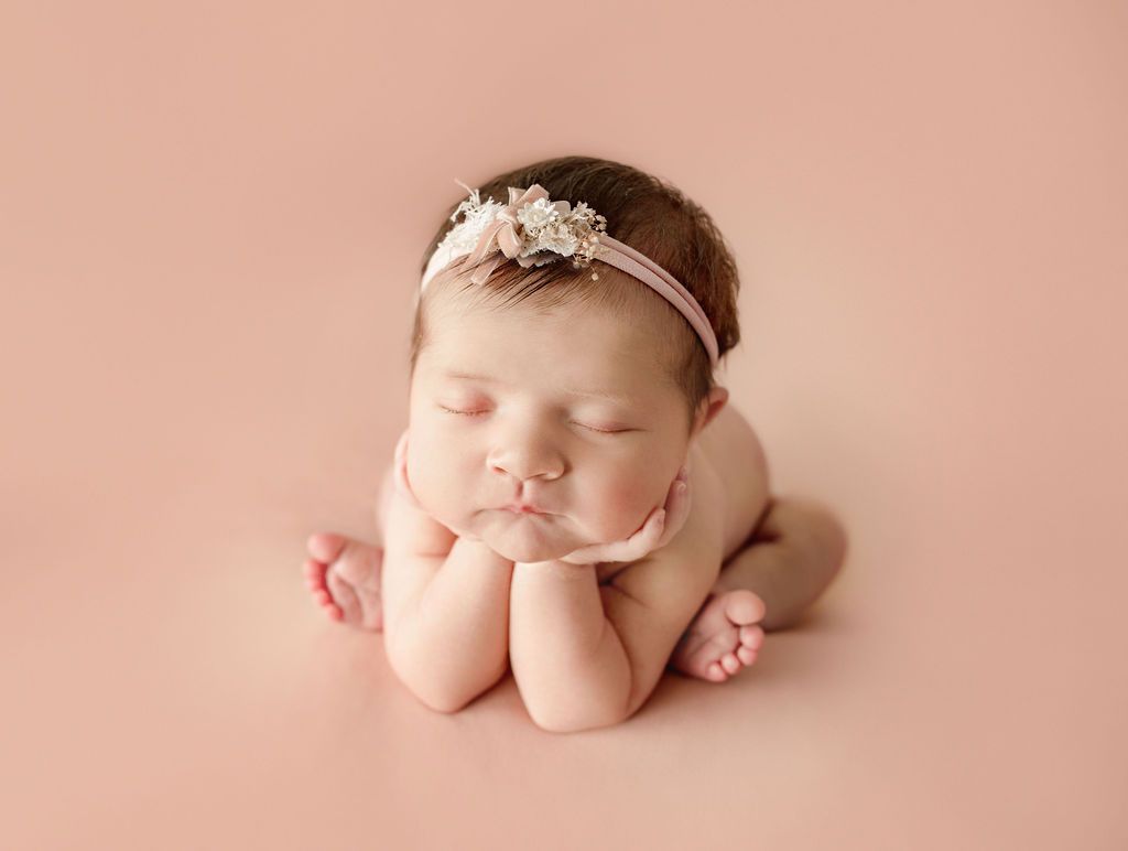 Newborn baby, eyes closed, resting chin on hands, wearing a floral headband, on a pink backdrop.