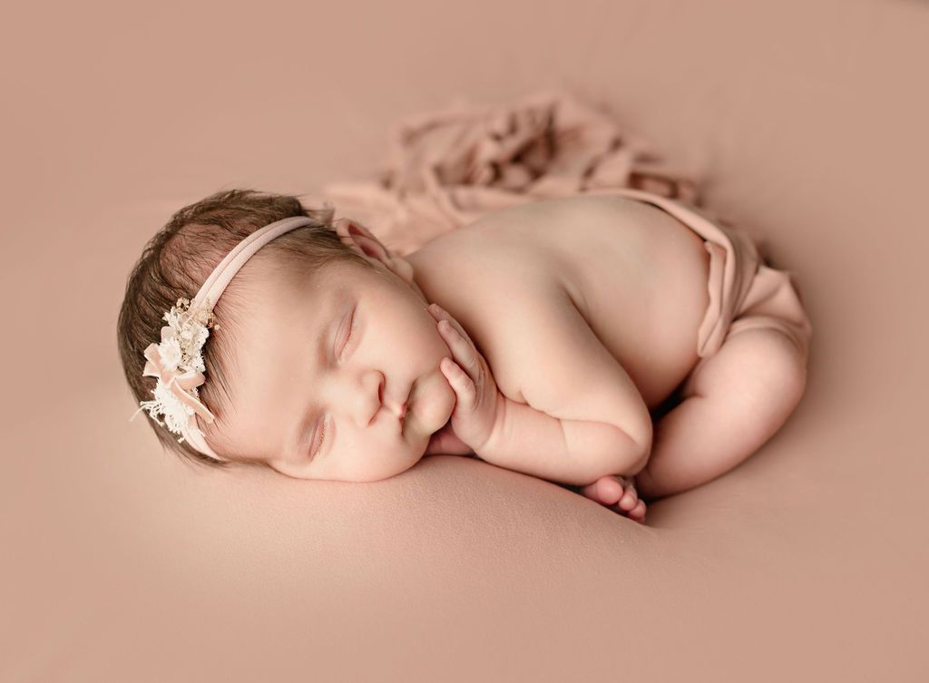 Newborn baby sleeping, wearing a floral headband and nude-colored diaper, resting on peach fabric.