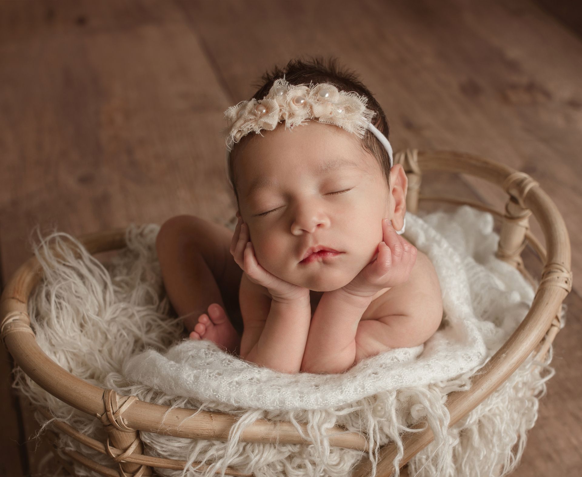 Newborn resting in a woven basket, resting head on hands; wearing a floral headband.