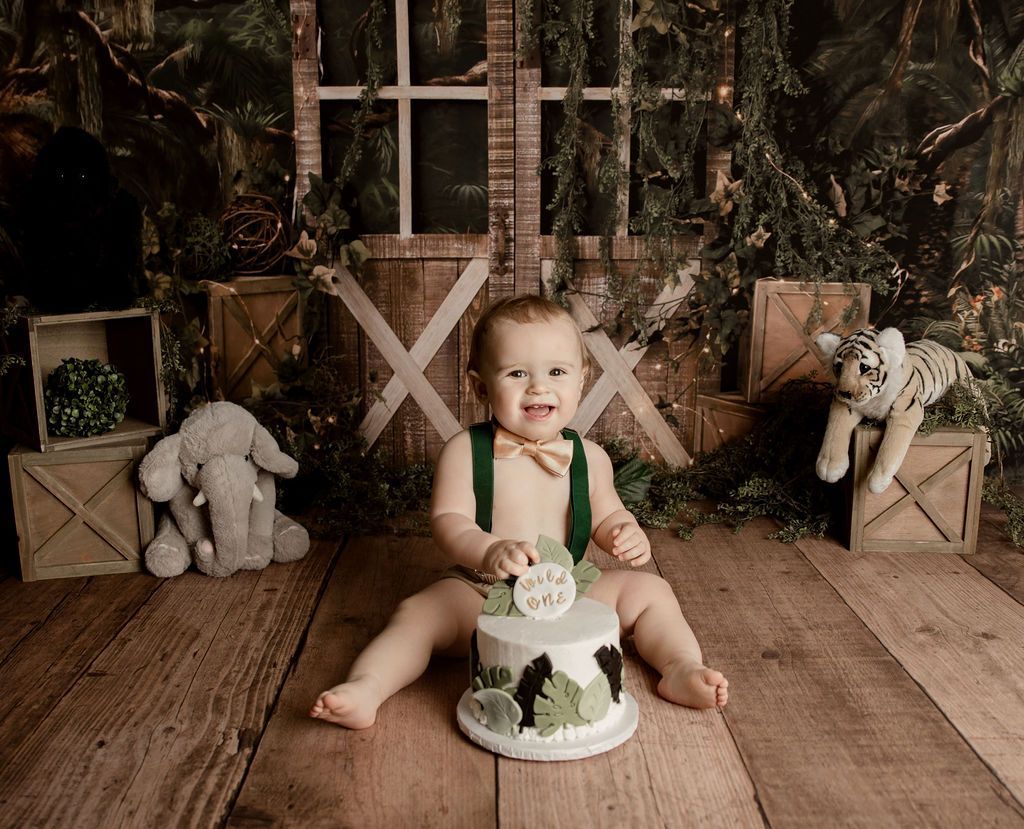 Baby sitting in front of a cake, wearing suspenders and bow tie. Jungle-themed background with stuffed animals.