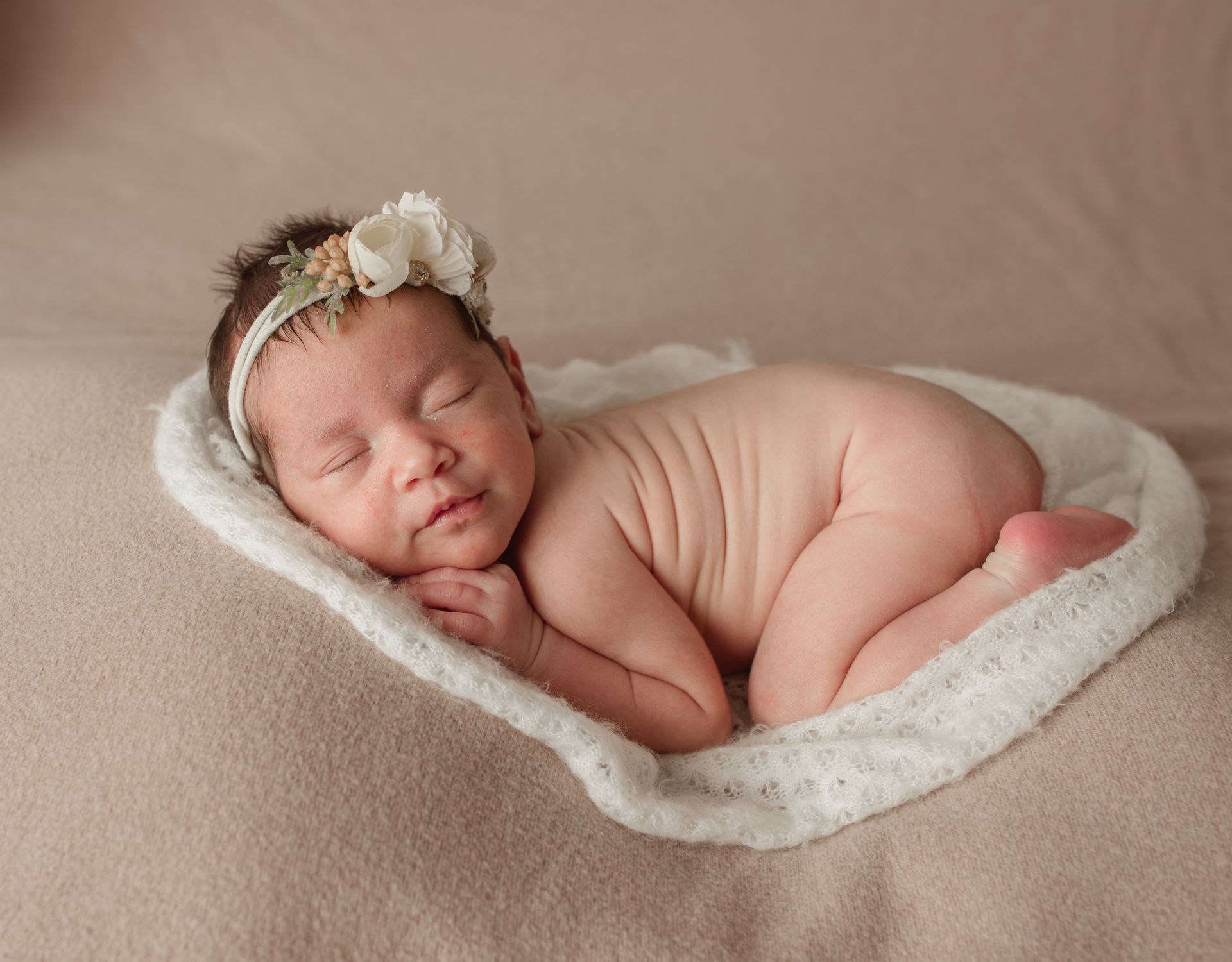 Newborn baby sleeping on a white, heart-shaped blanket, wearing a floral headband.