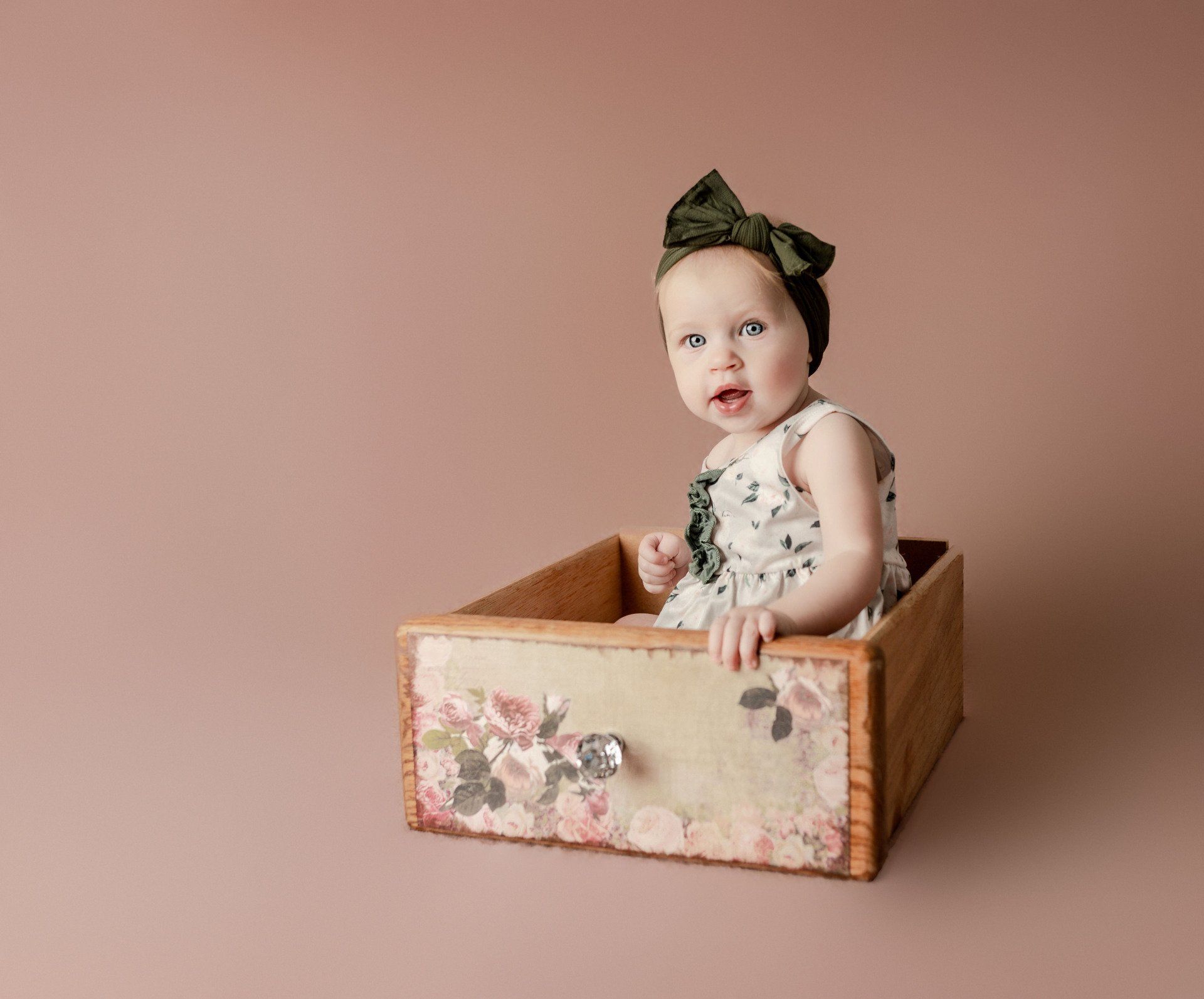 Baby in a floral dress sits in a wooden drawer, wearing a green bow. Background is dusty rose.