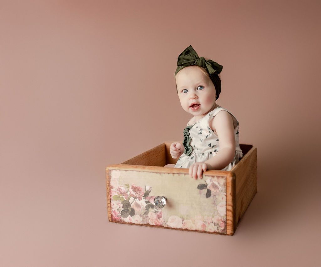Baby in floral outfit sits in a decorated wooden drawer, looking forward with a surprised expression.