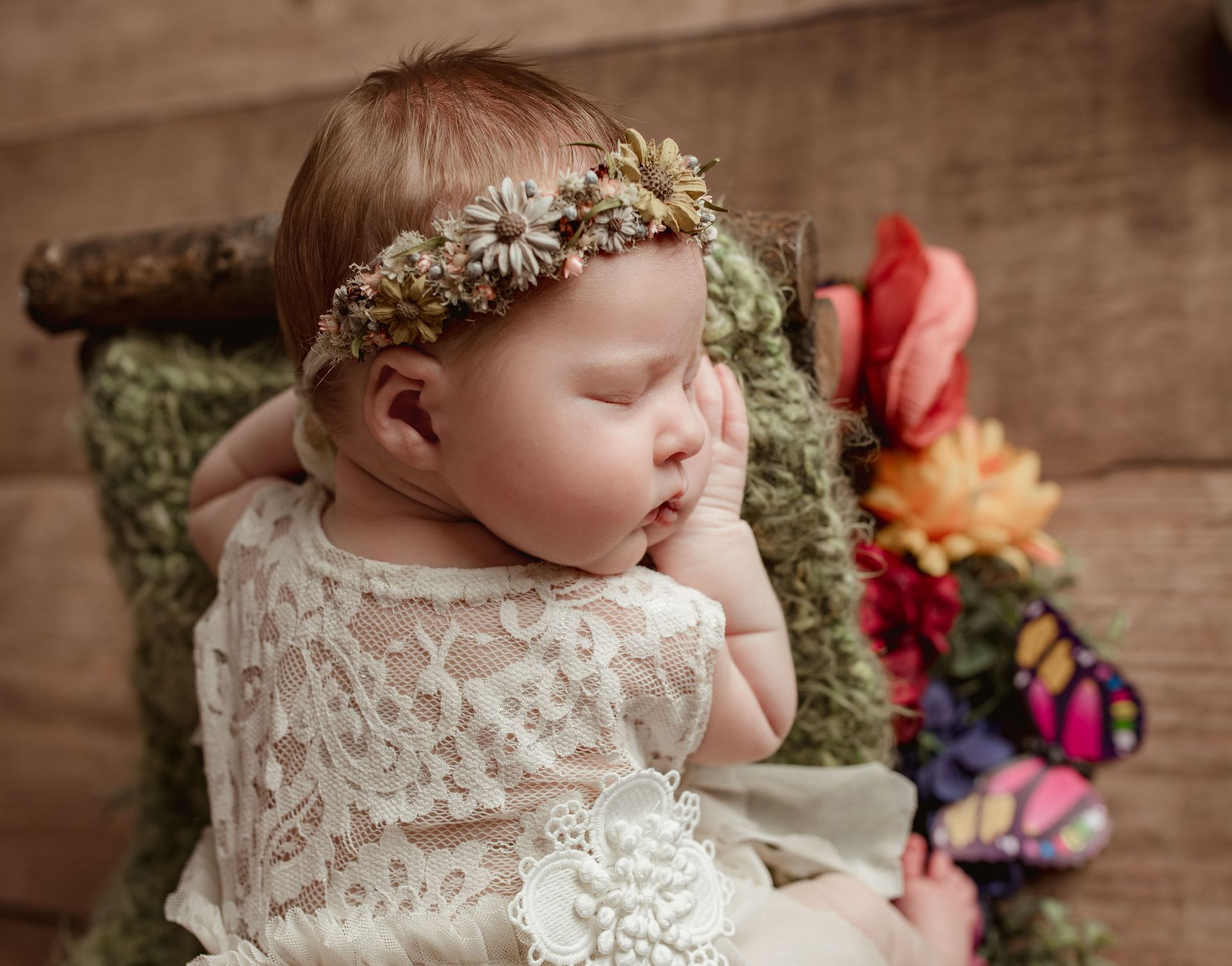 Sleeping baby in white lace dress and flower crown, resting on green blanket with floral accents.