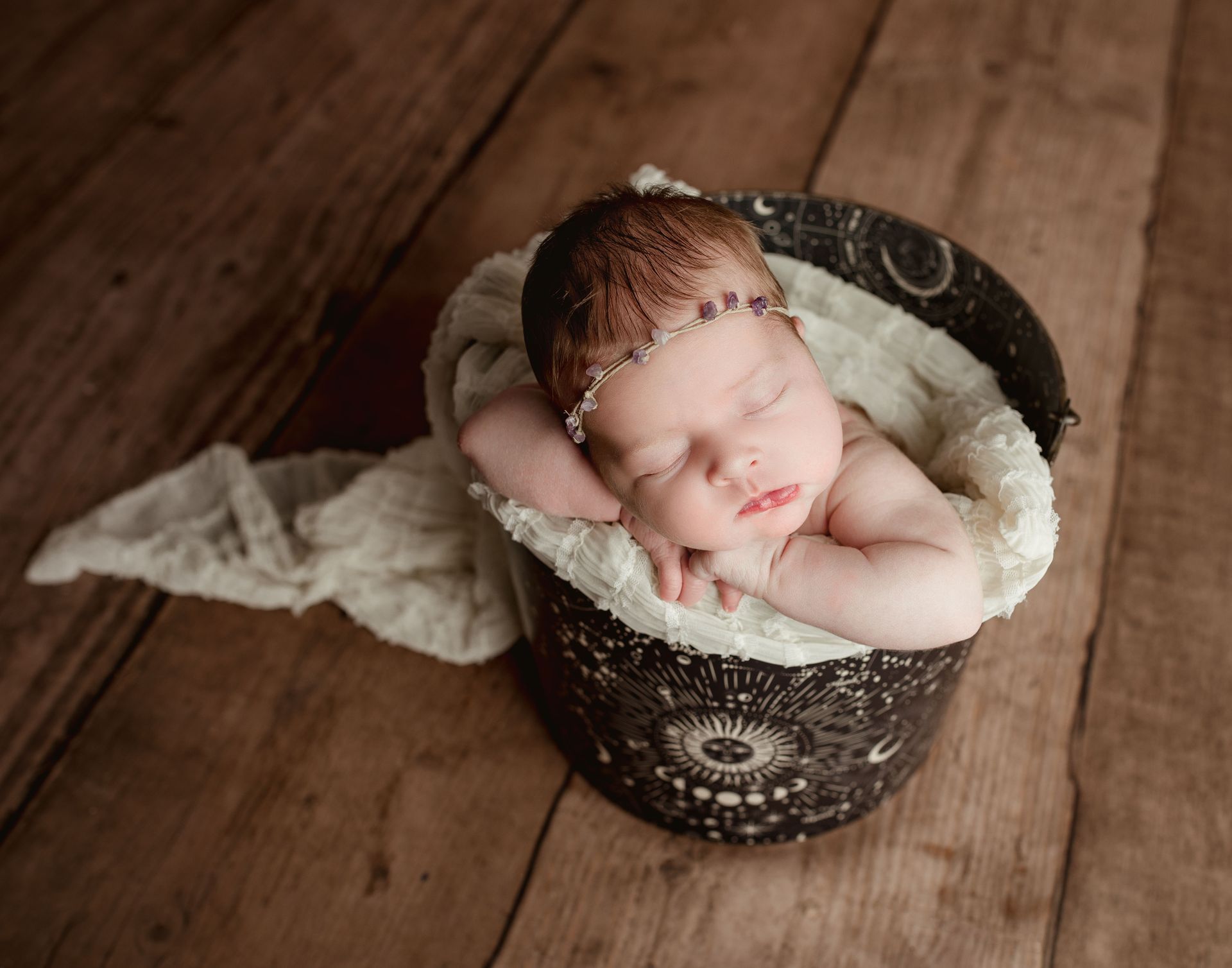 Newborn baby sleeping in a decorative bucket, resting on arms. White blanket, wooden background.