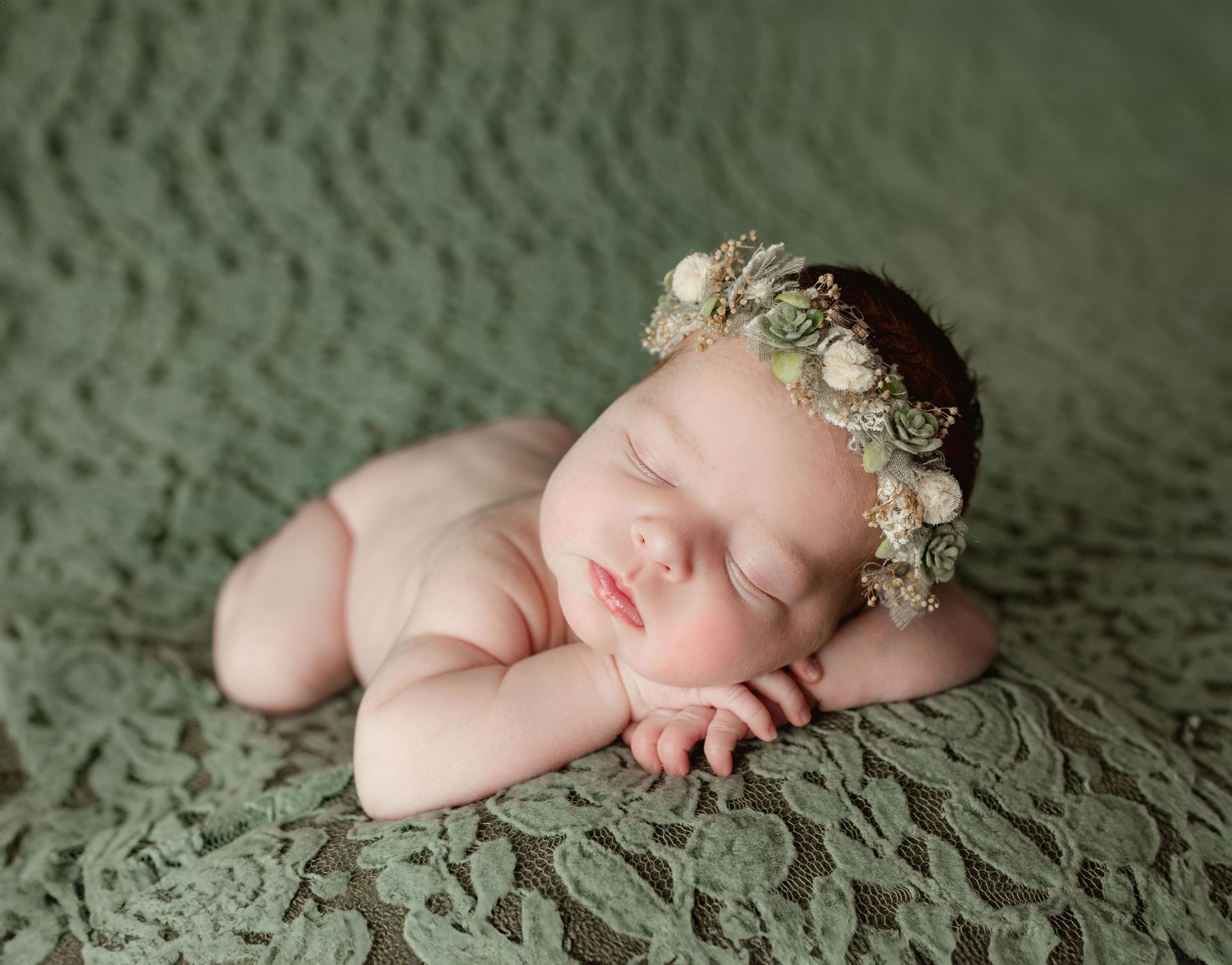 Newborn baby sleeping, wearing floral crown, on green lace blanket.