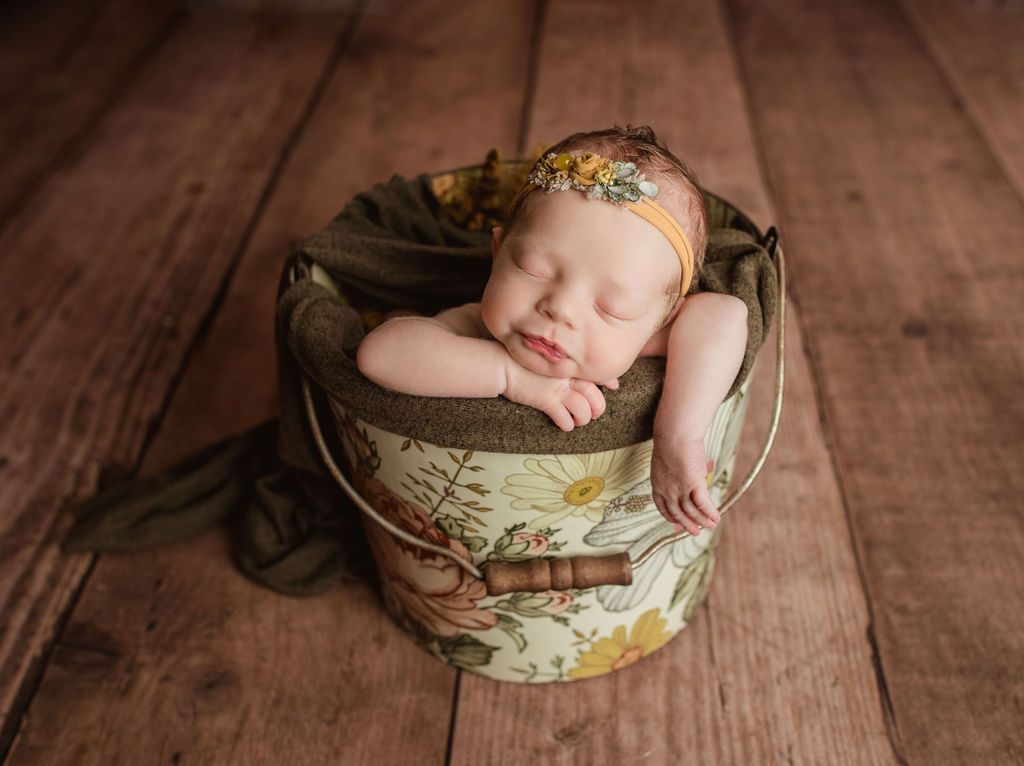 Newborn baby sleeping in a floral bucket, on a wood floor, wearing a yellow floral headband.