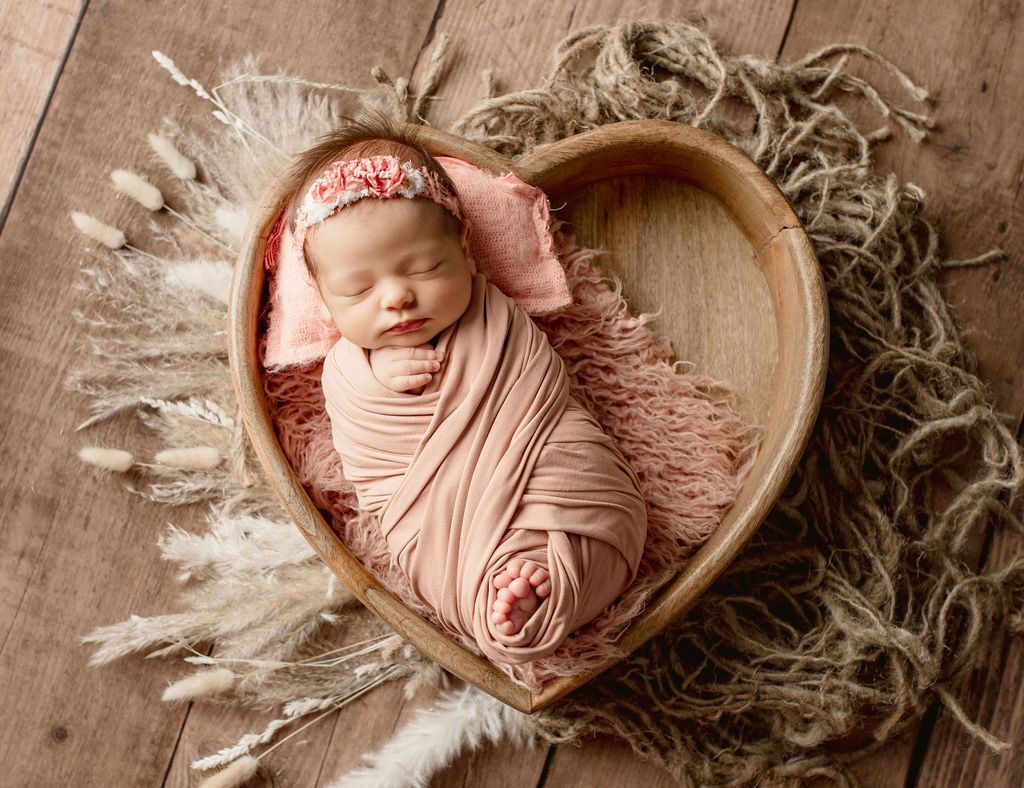 Newborn wrapped in pink blanket, in heart-shaped wooden bowl, on wood surface.
