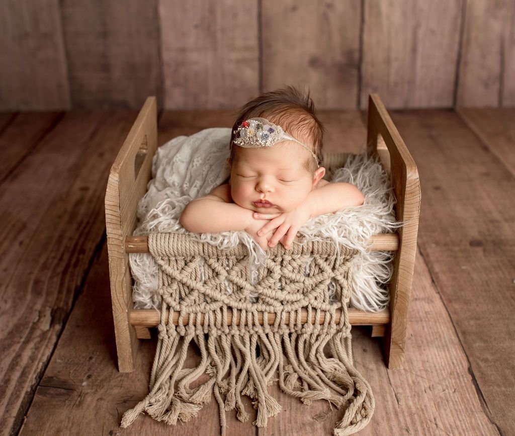 Newborn asleep in a small wooden bed with a macrame headboard, wearing a floral headband, on a wooden floor.