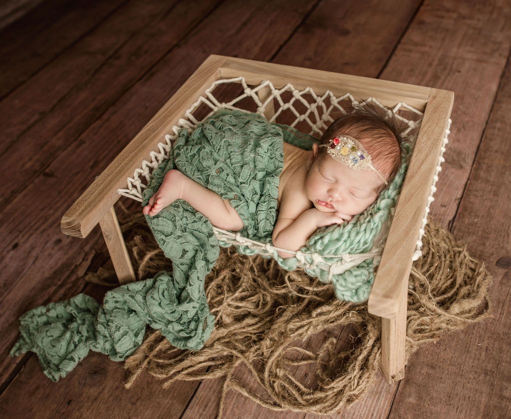 Newborn baby sleeping in a small wooden hammock, wrapped in a green blanket, on a brown wooden floor.