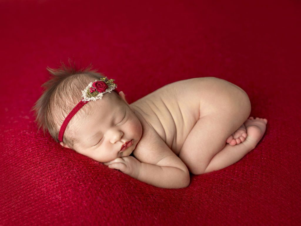 Newborn baby sleeping on red fabric, wearing a red floral headband.