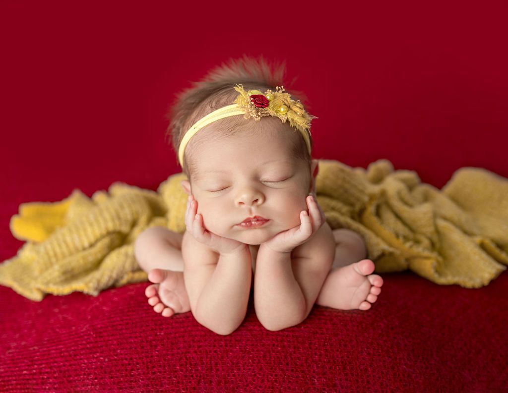 Newborn baby with eyes closed, resting on hands and wrapped in yellow blanket, on a red background.
