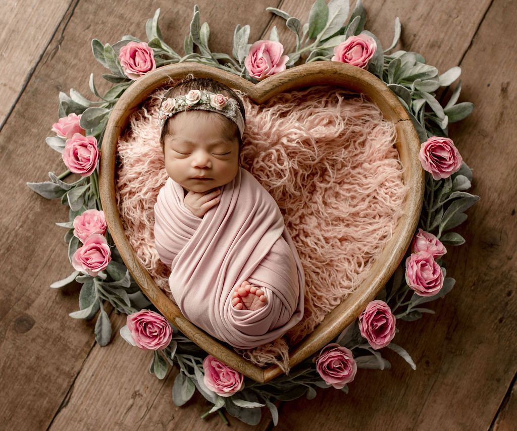 Newborn swaddled in pink, in a heart-shaped wooden basket with pink roses and greenery on a wood surface.