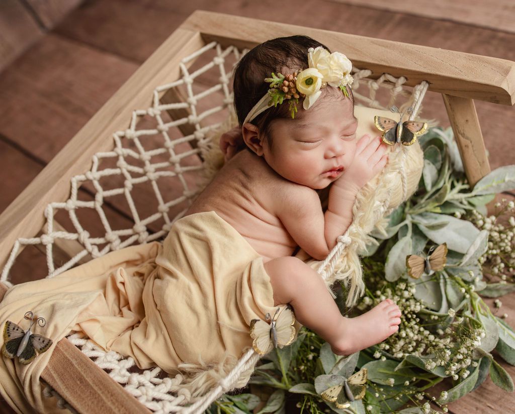 Newborn in beige fabric and flower crown, sleeping in a wooden hammock decorated with greenery and butterflies.