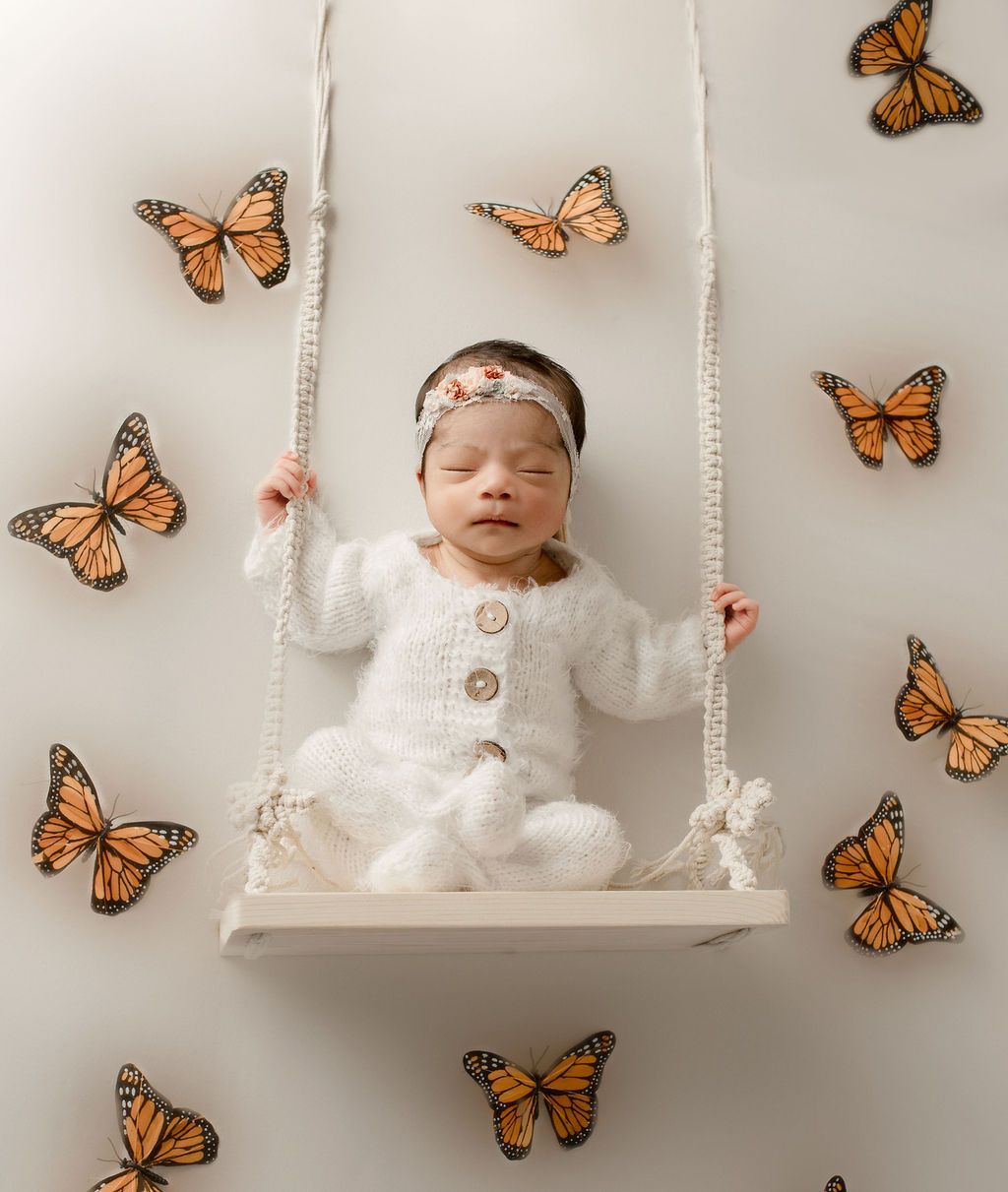 Newborn on a swing, surrounded by butterfly decorations, wearing a white knit outfit and headband.