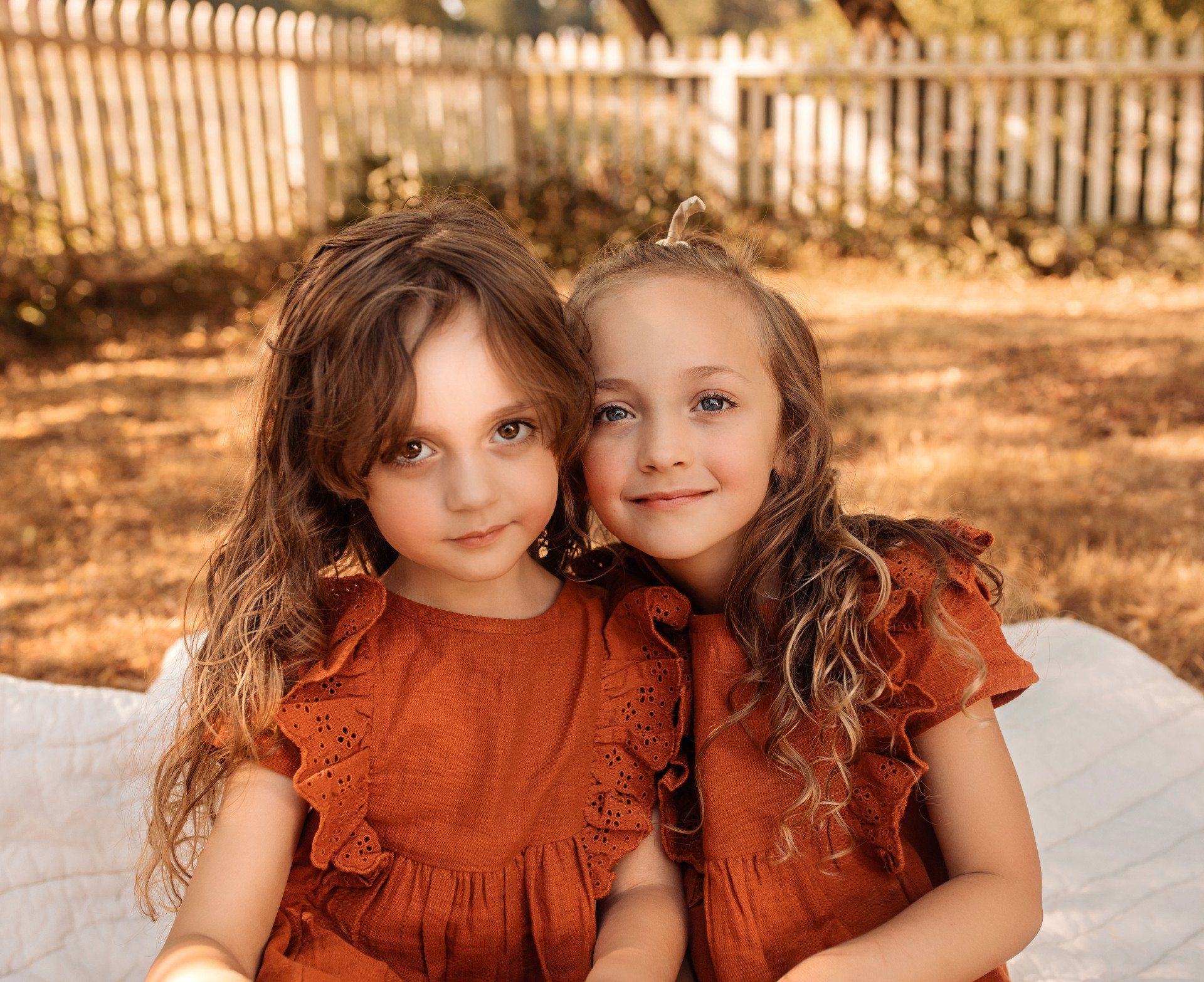 Two young girls in rust-colored dresses smile. They are seated, with a white fence and foliage background.