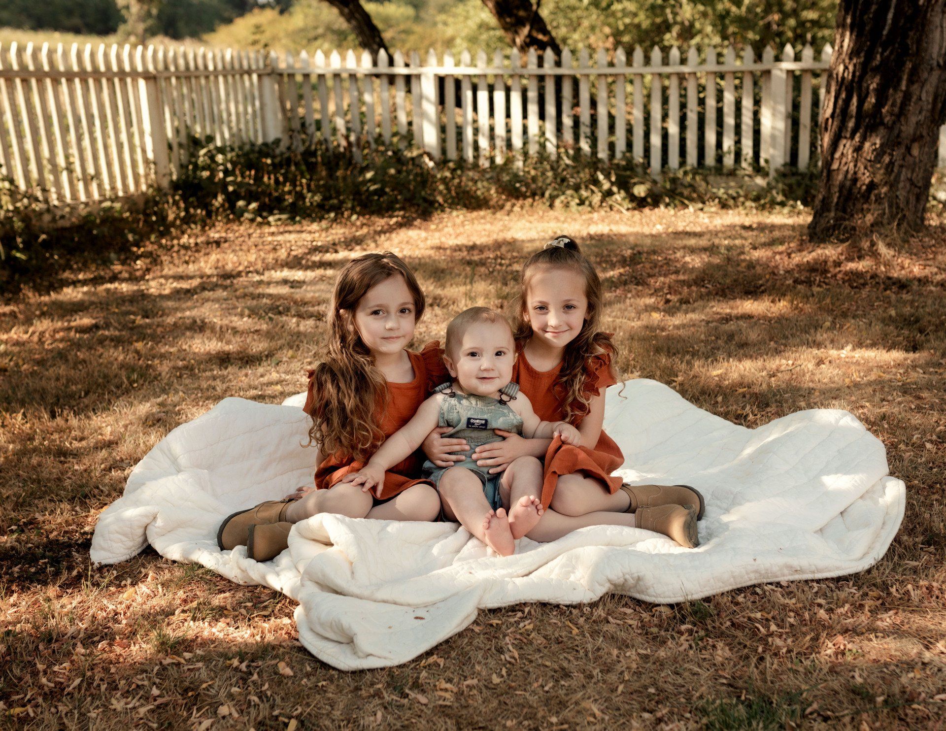 Three children sit on a blanket outdoors in front of a white picket fence.