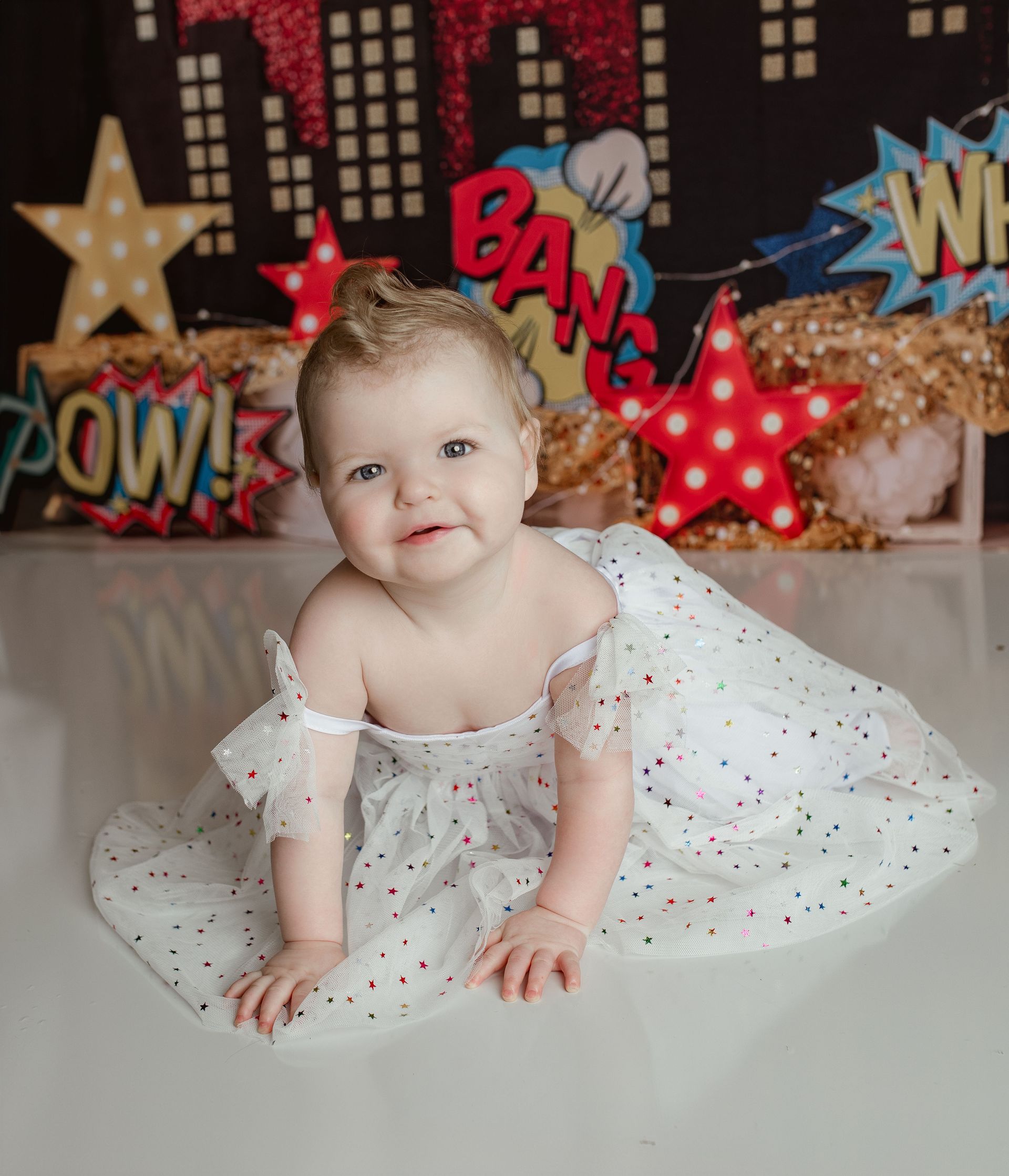 Smiling toddler in white dress with red dots, superhero-themed background.
