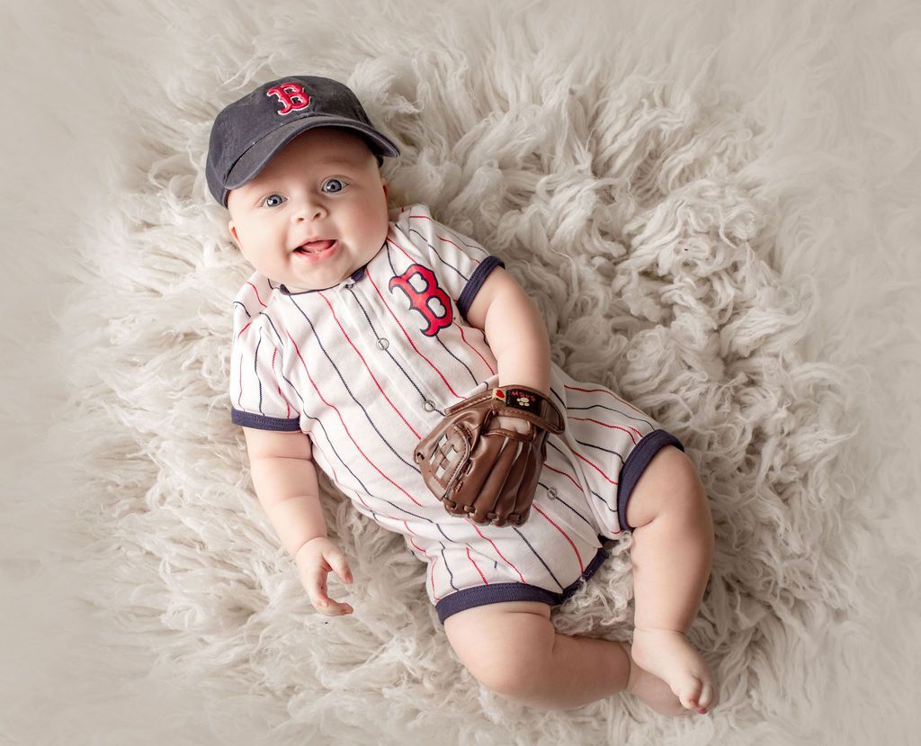 Baby in Red Sox baseball outfit and cap smiles, holding a glove, on a fuzzy white surface.
