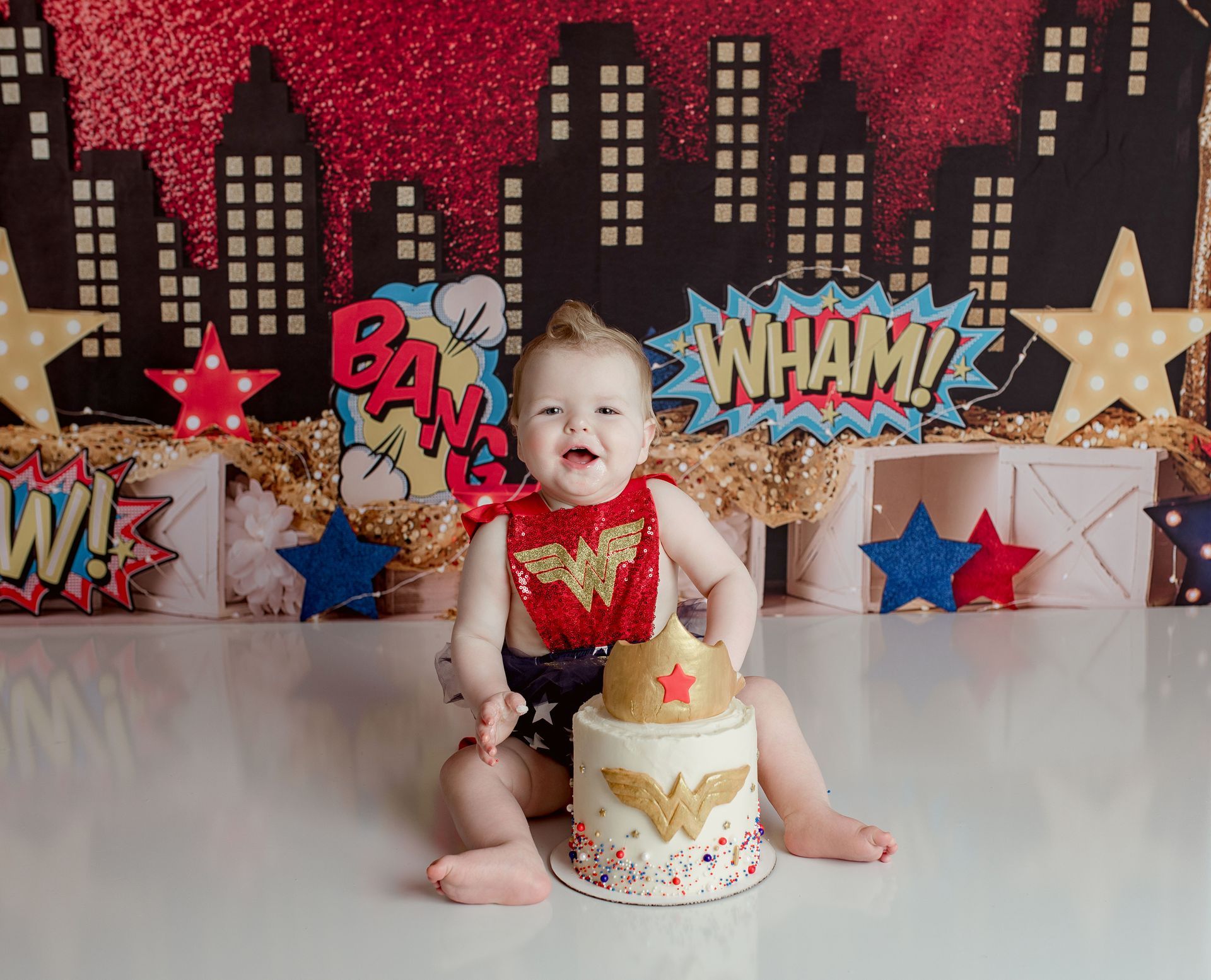 Baby in Wonder Woman attire, smiling, with cake and comic book backdrop.