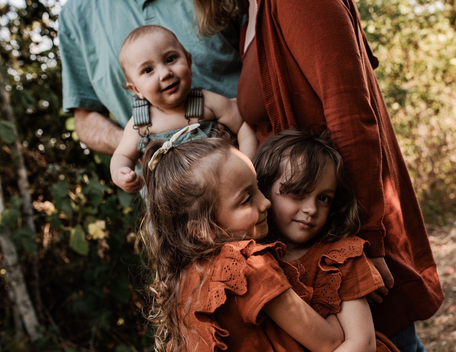 Family portrait in outdoor setting. Two young children hugging, a baby peering over, adults partially visible.