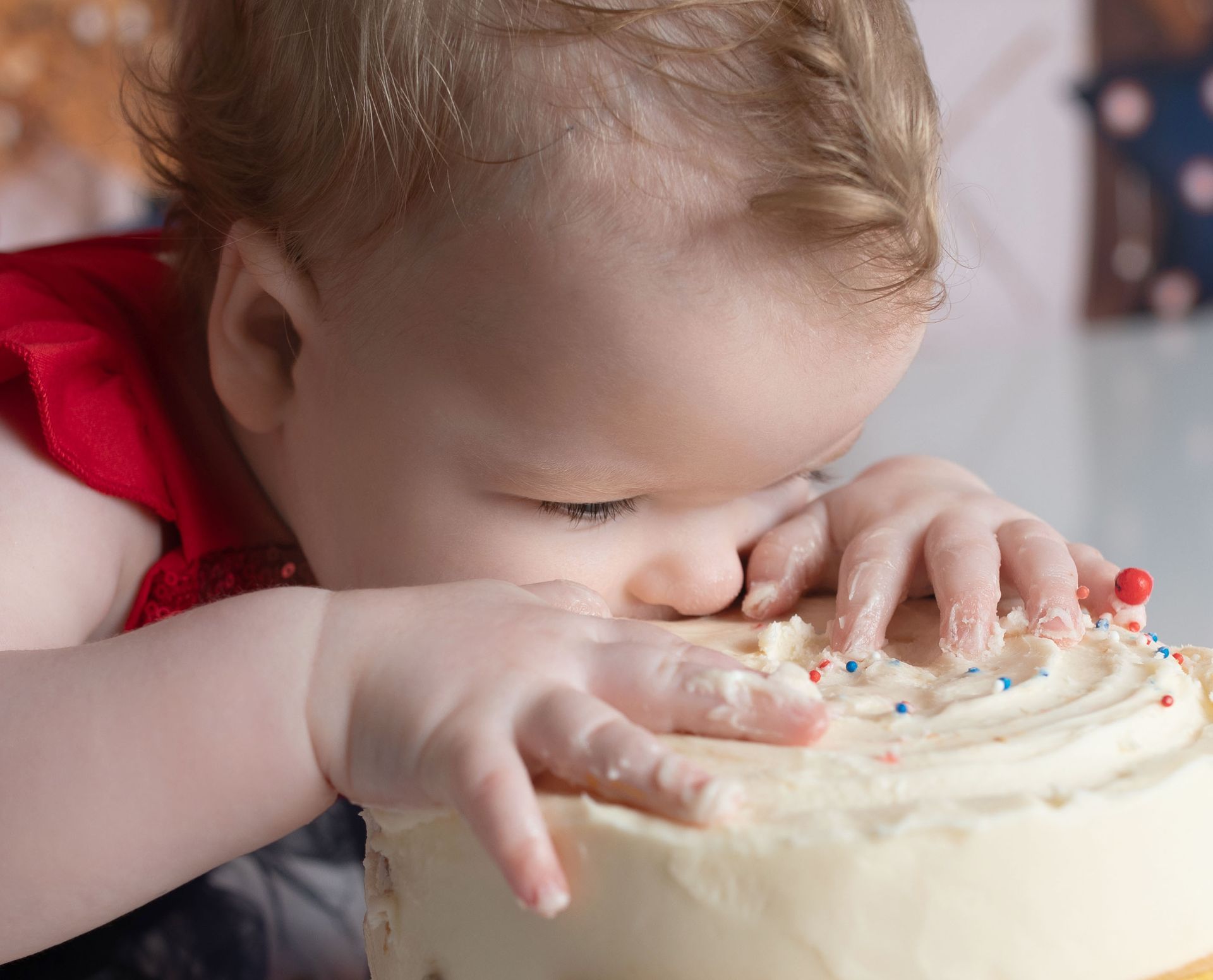 Baby eating a cake with hands, white frosting, and colorful sprinkles. Red shirt, blurred background.
