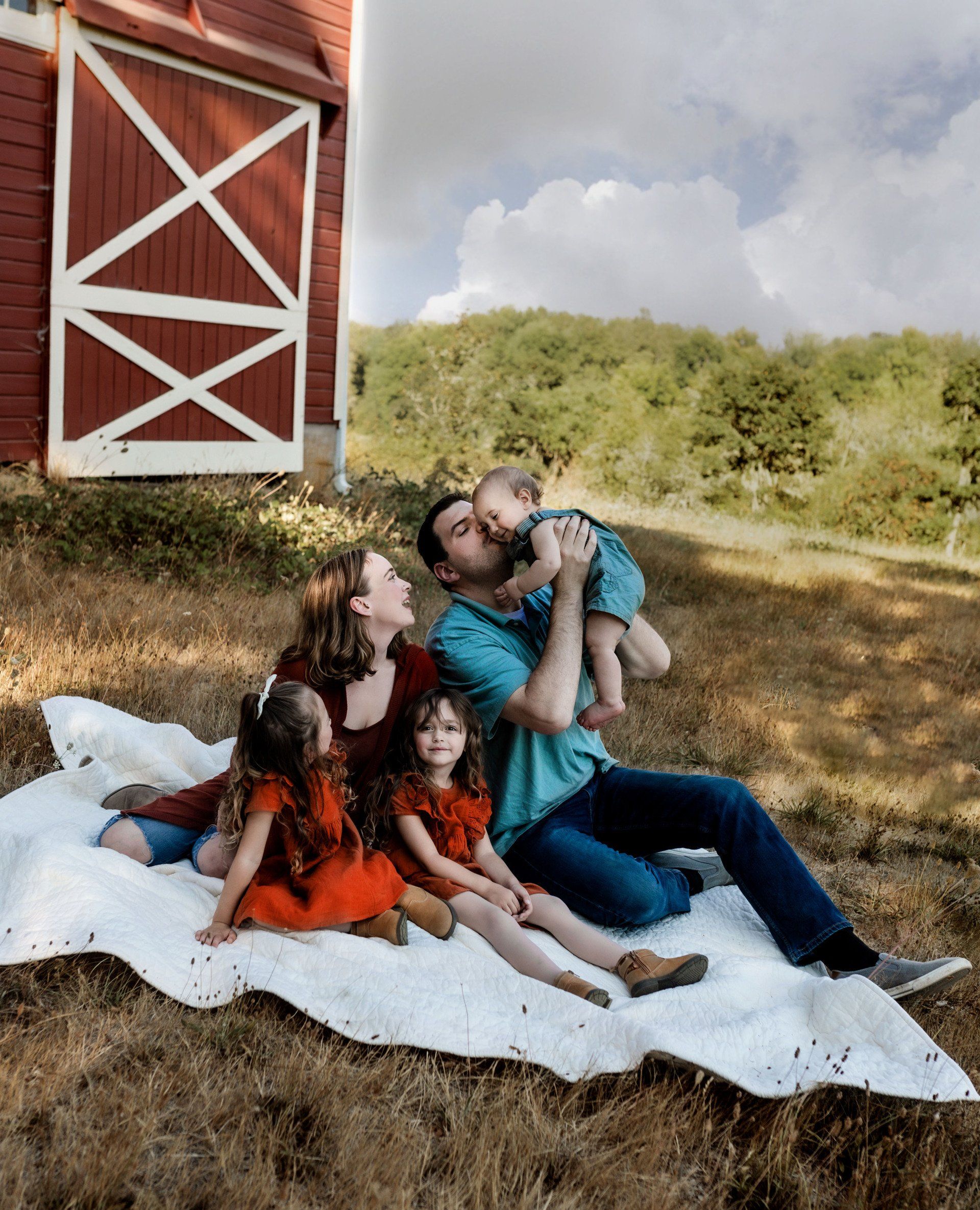 Family of five on a blanket near a red barn; adults kissing baby, girls sitting.