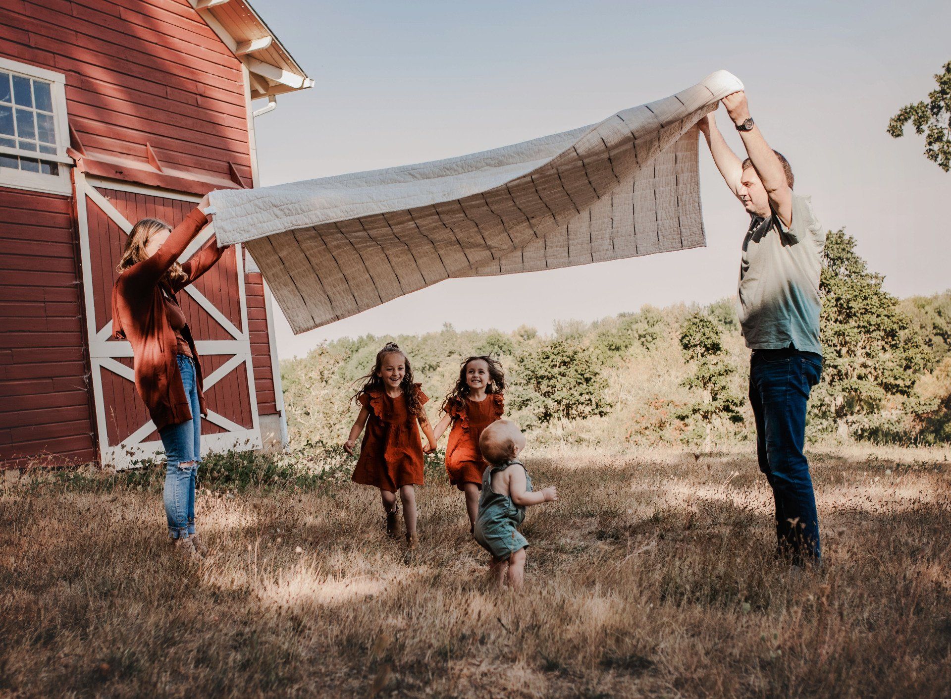 Family holding up a patterned blanket in a field near a red barn; children run toward the blanket.