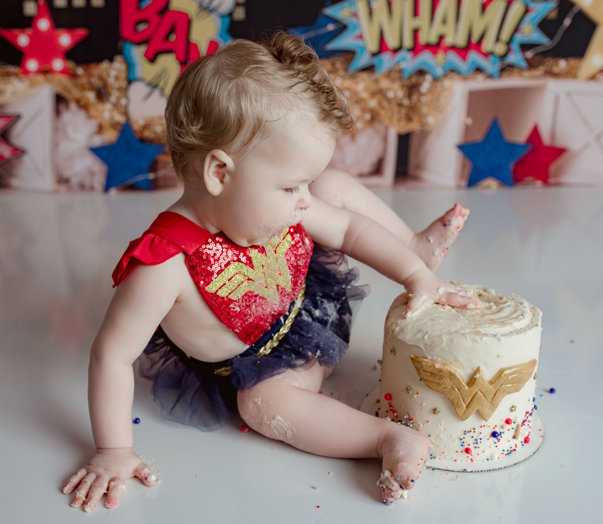 Baby in Wonder Woman outfit with a cake, smearing frosting. Red, blue, gold colors; birthday backdrop.