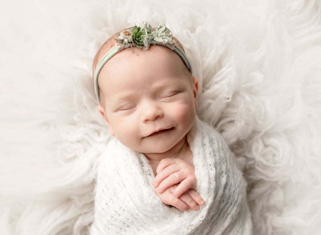 Newborn wrapped in a white blanket, wearing a floral headband, smiling.