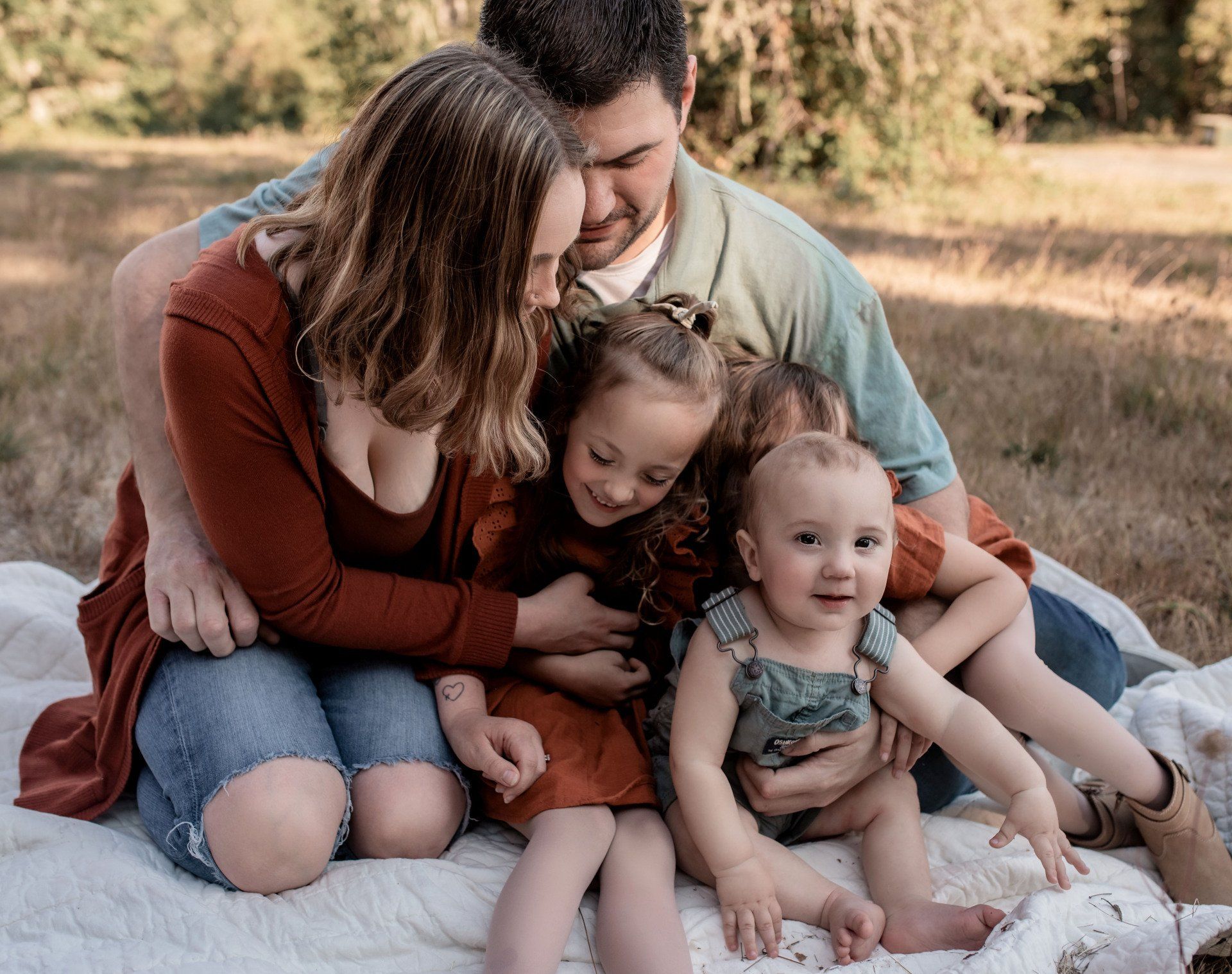 Family of five embraces on a blanket outdoors; parents hug children, smiling.