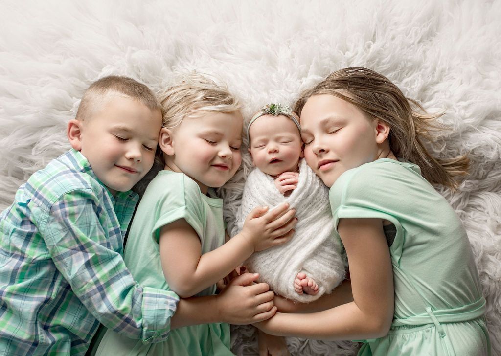 Four children gently embrace a swaddled newborn on a soft white blanket.