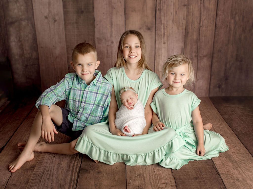 Four children seated on wooden floor, newborn swaddled in middle. All are wearing mint green or teal.