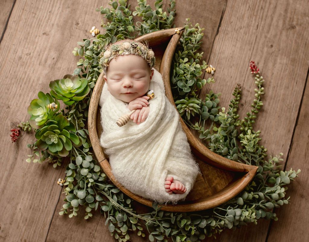 Newborn swaddled in cream-colored blanket, nestled in a wooden moon-shaped bowl surrounded by greenery, on a wooden surface.