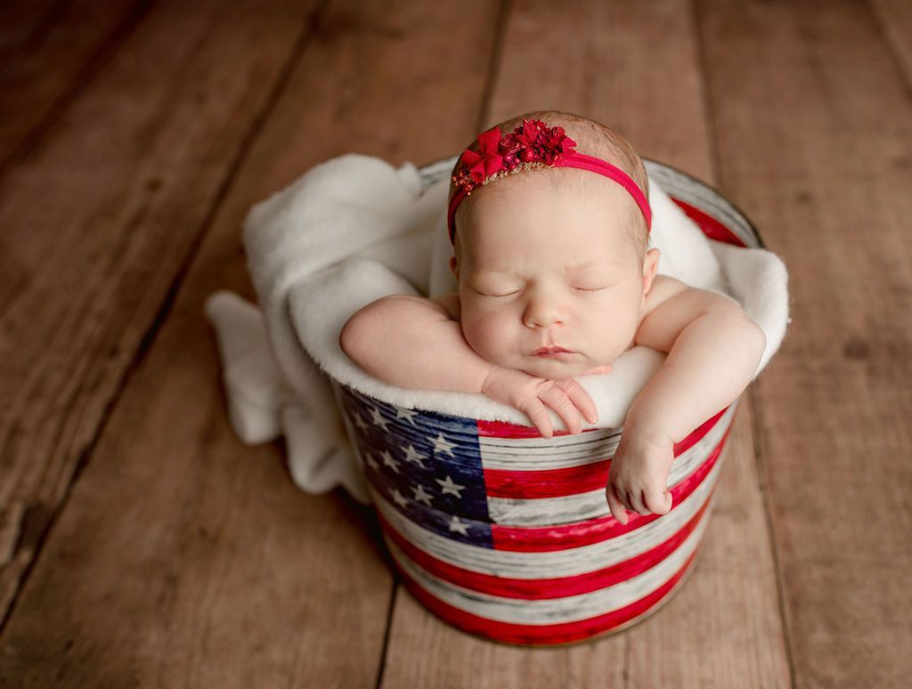 Baby sleeping in a patriotic bucket with a red headband, on a wood surface.