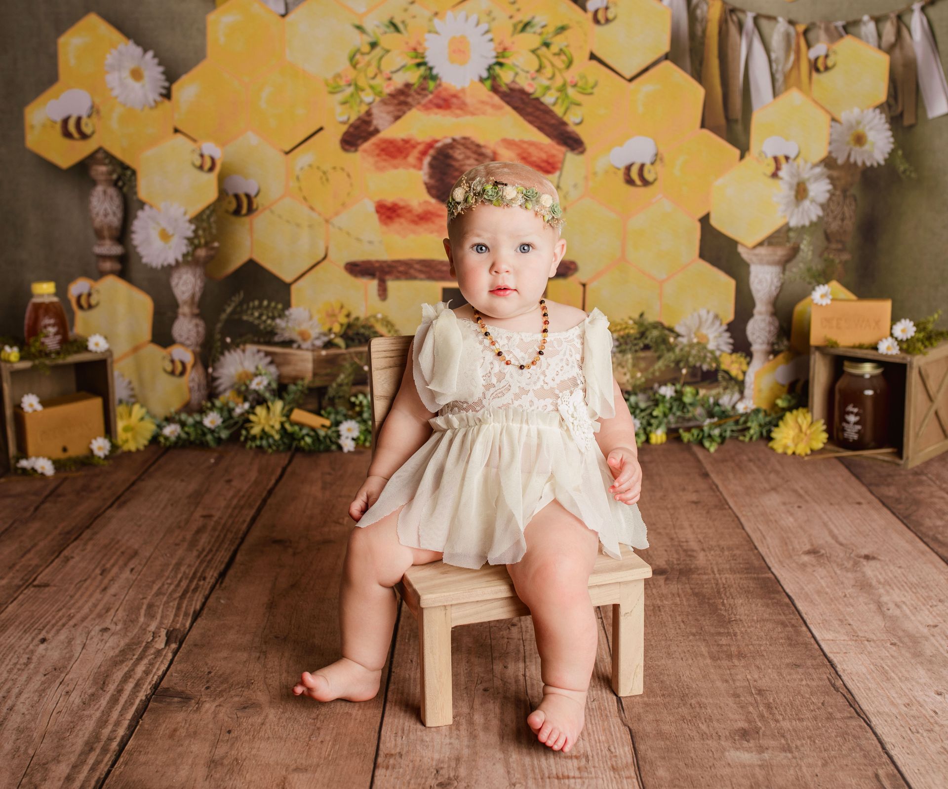 Baby in a cream outfit sits on a small chair in front of a bee-themed backdrop, with flowers and honey decorations.