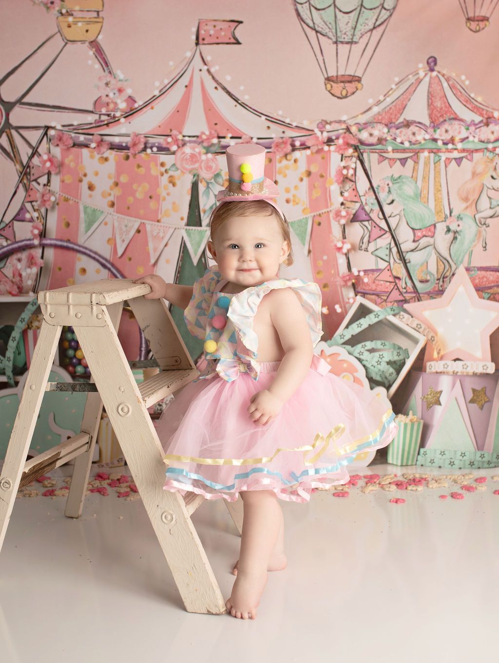 Baby in circus-themed outfit, smiling, posing near a wooden ladder, colorful backdrop.
