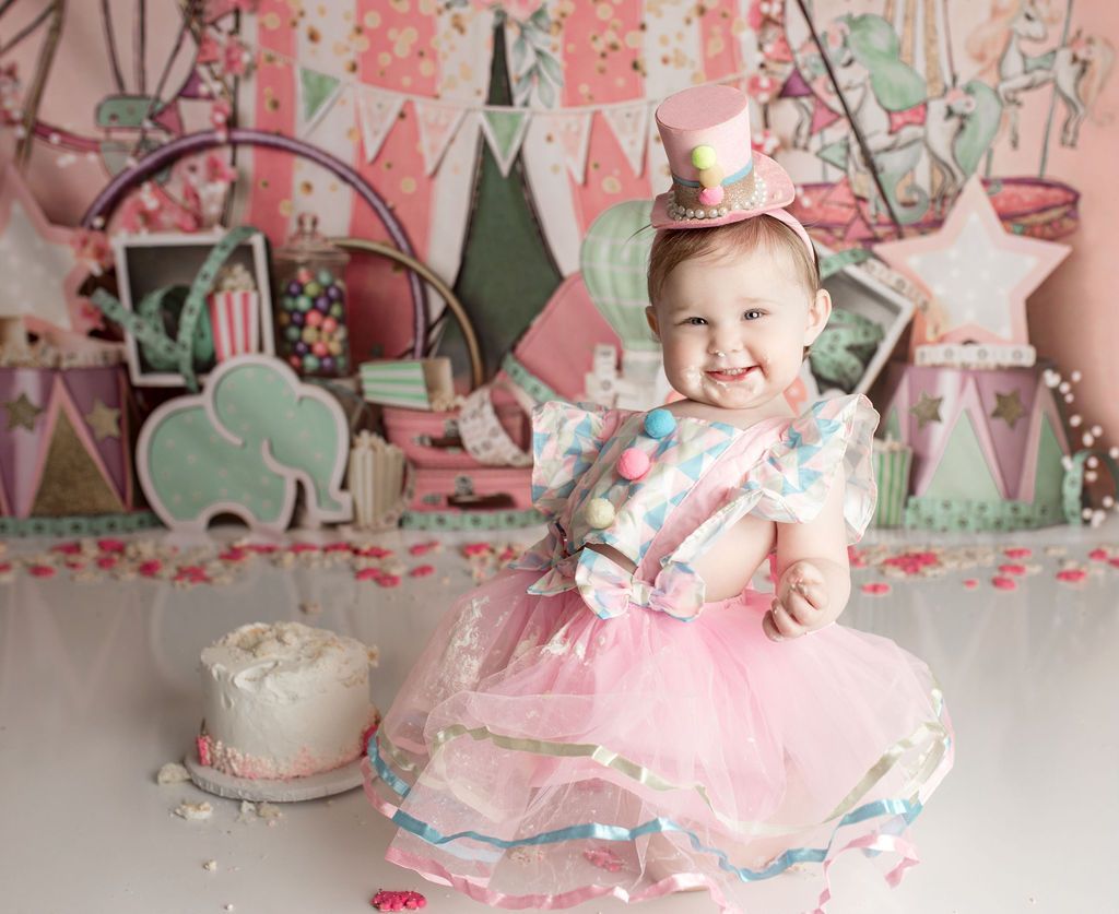 Smiling baby in circus-themed outfit, celebrating a birthday, with cake and backdrop.